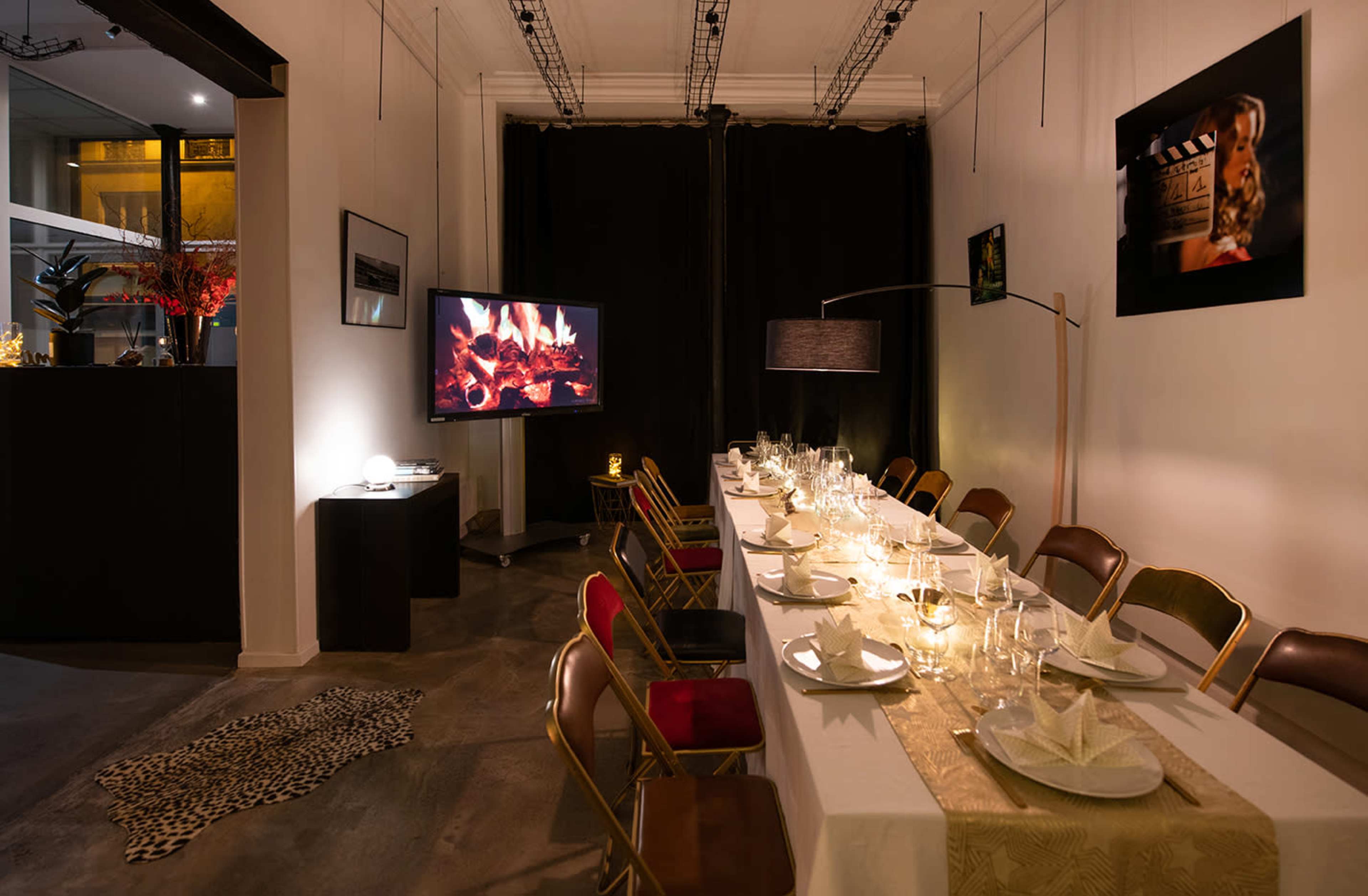 The scene shows a dining area set with a long table adorned with plates and glasses, flanked by chairs, and featuring a television displaying a fireplace backdrop.