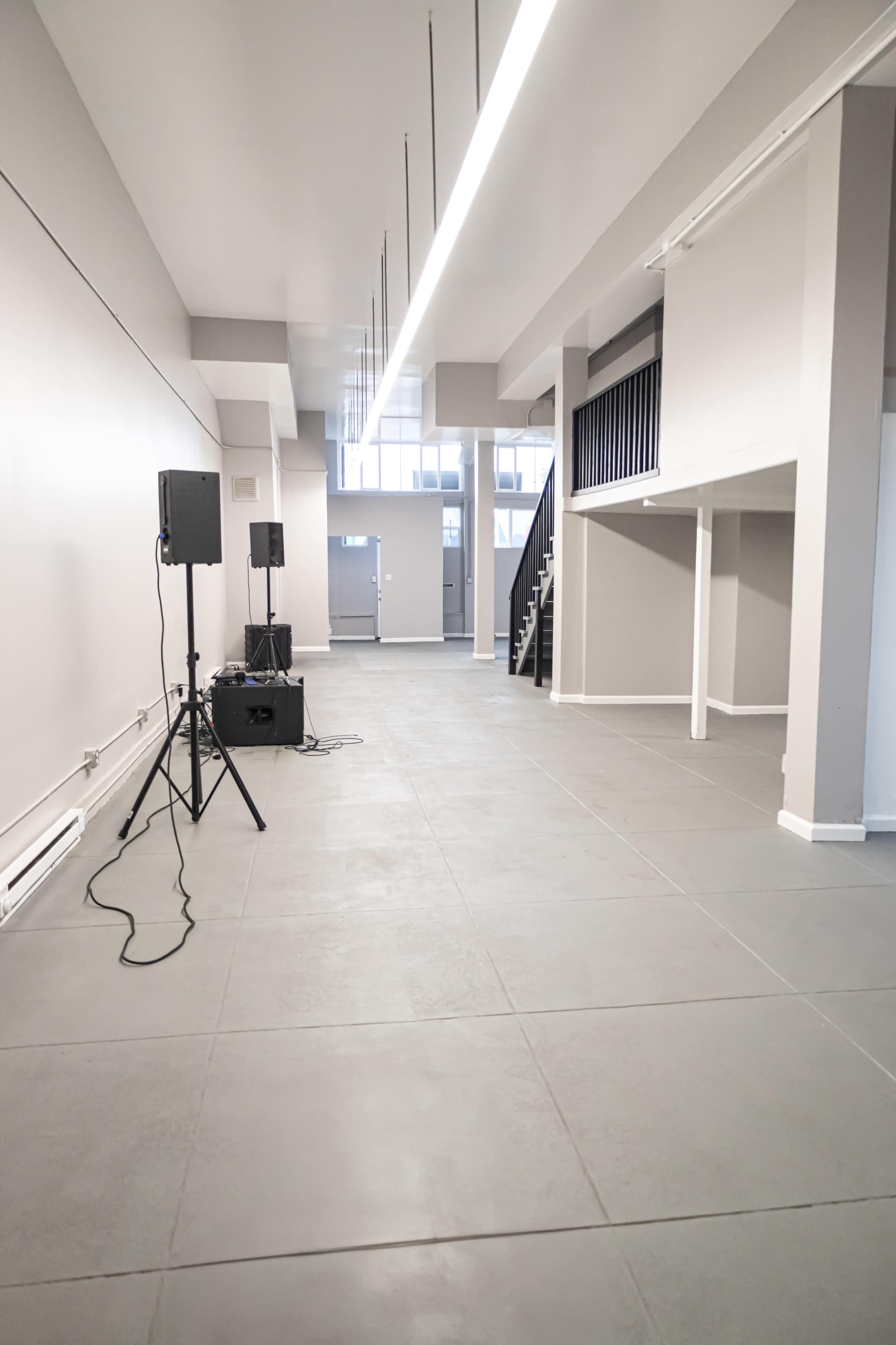 The image shows a spacious, empty interior with gray tile flooring, large windows, and a staircase in the background along with audio equipment on the left side.