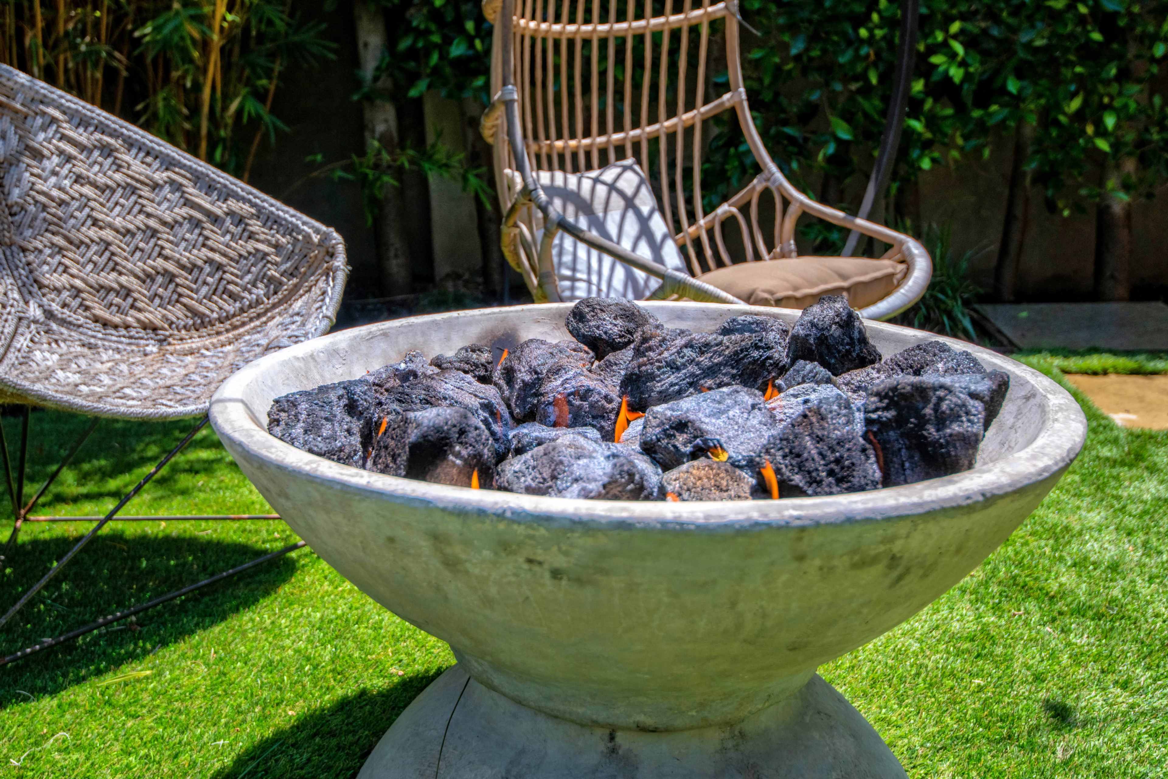 A round fire pit filled with black rocks is centrally positioned on a grassy area next to a woven chair.