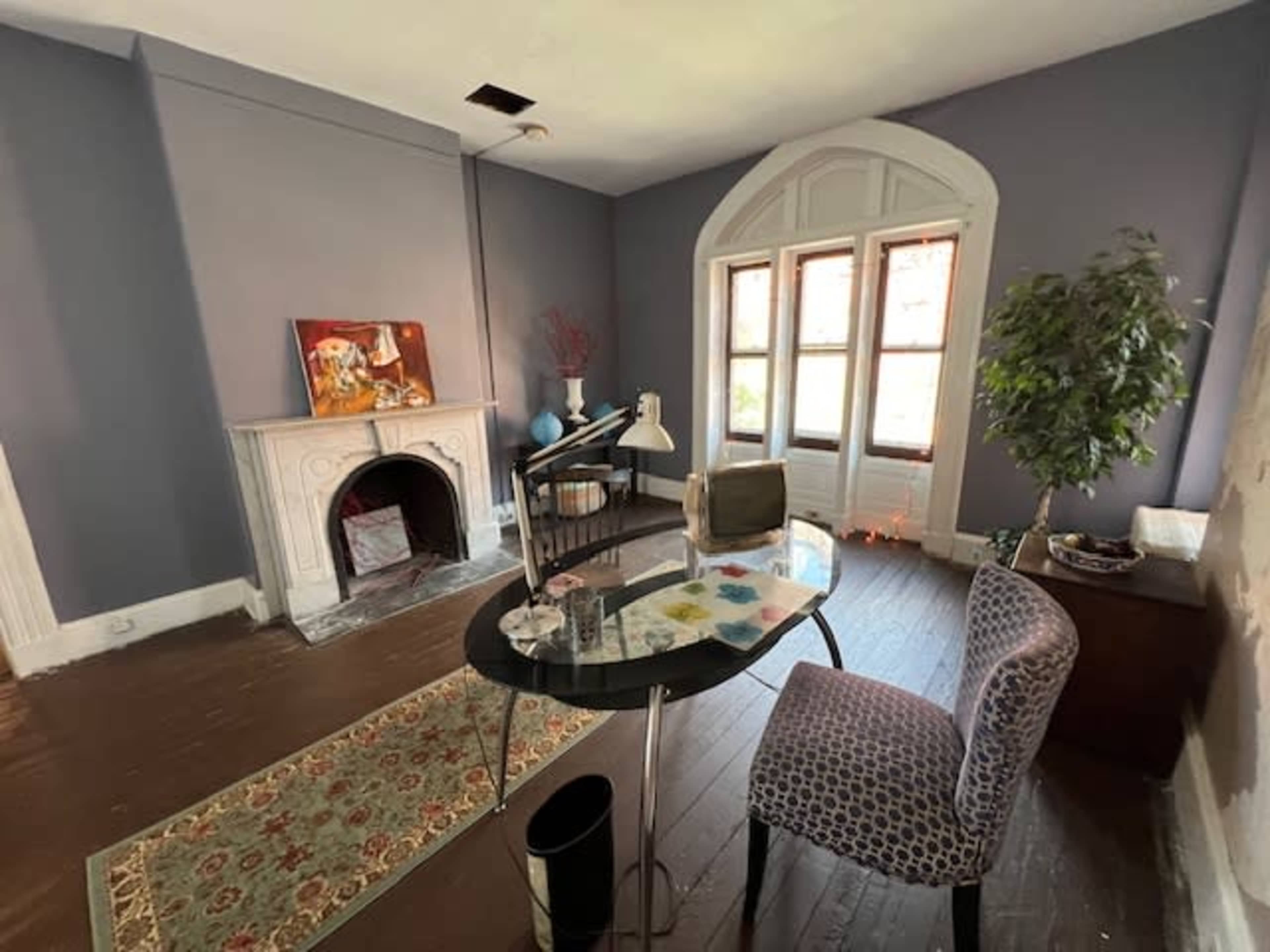 A room with a desk and chair, a fireplace, and large windows allowing natural light, featuring a potted plant and artwork on the wall.