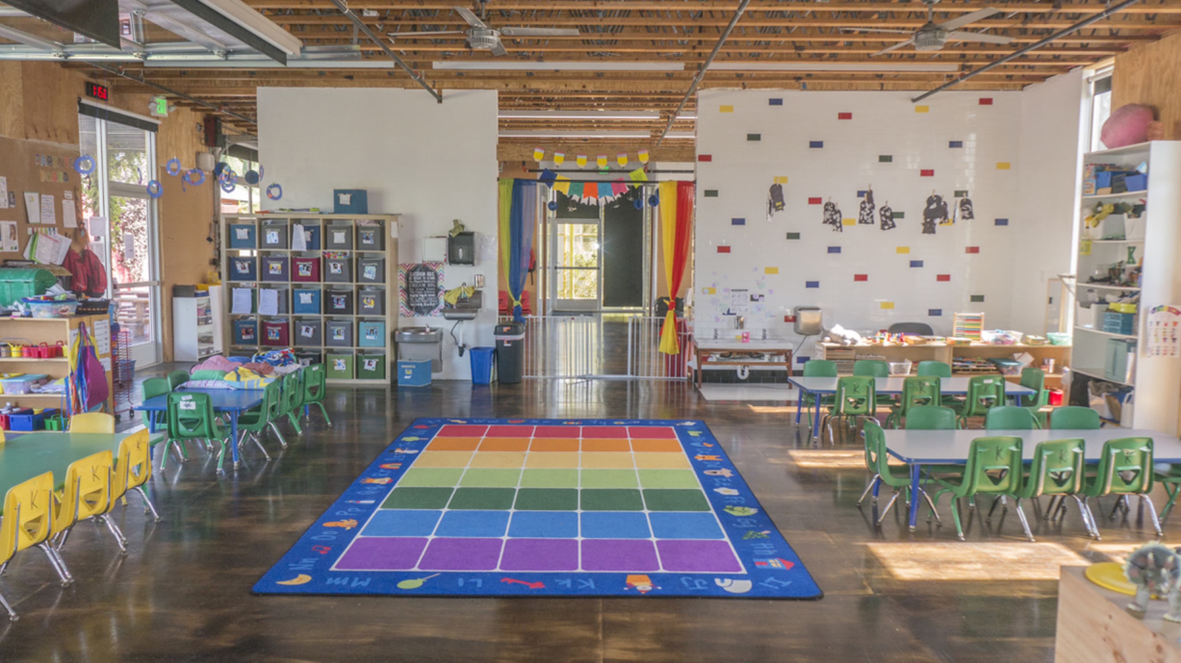A brightly lit classroom with colorful rugs, tables and chairs arranged for children, and a variety of educational materials on display.