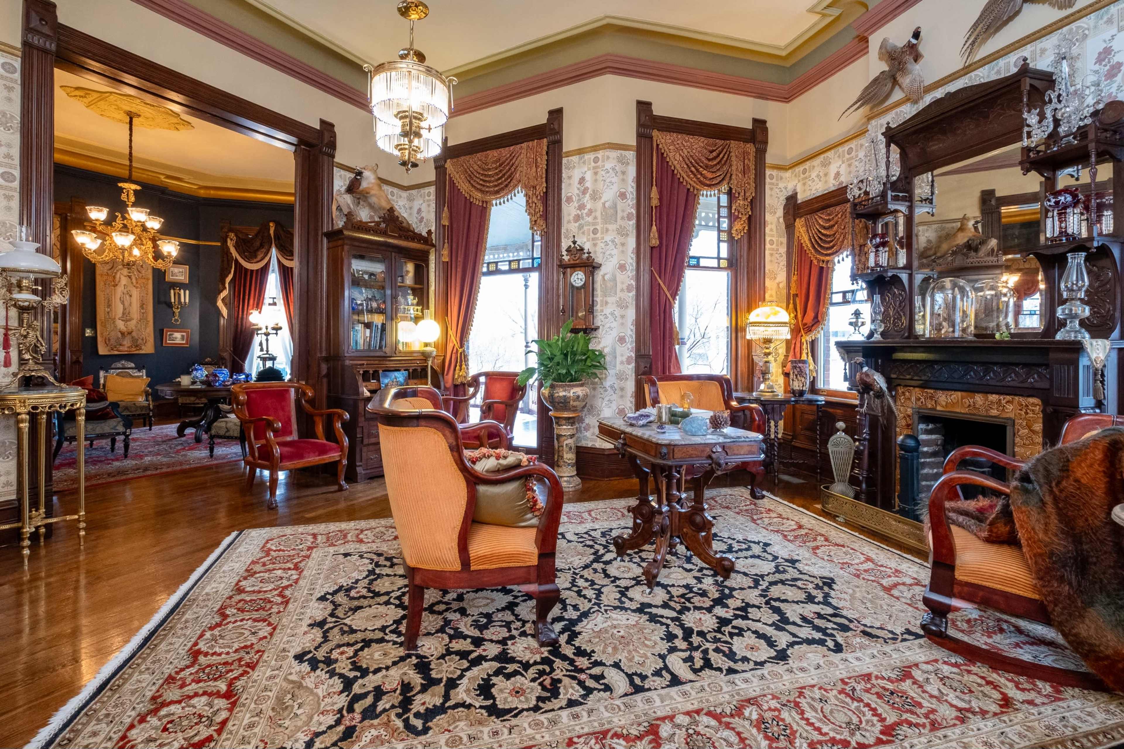 The image depicts an elegantly furnished sitting room with ornate decor, a patterned rug, and vintage furniture against a backdrop of floral wallpaper and large windows.
