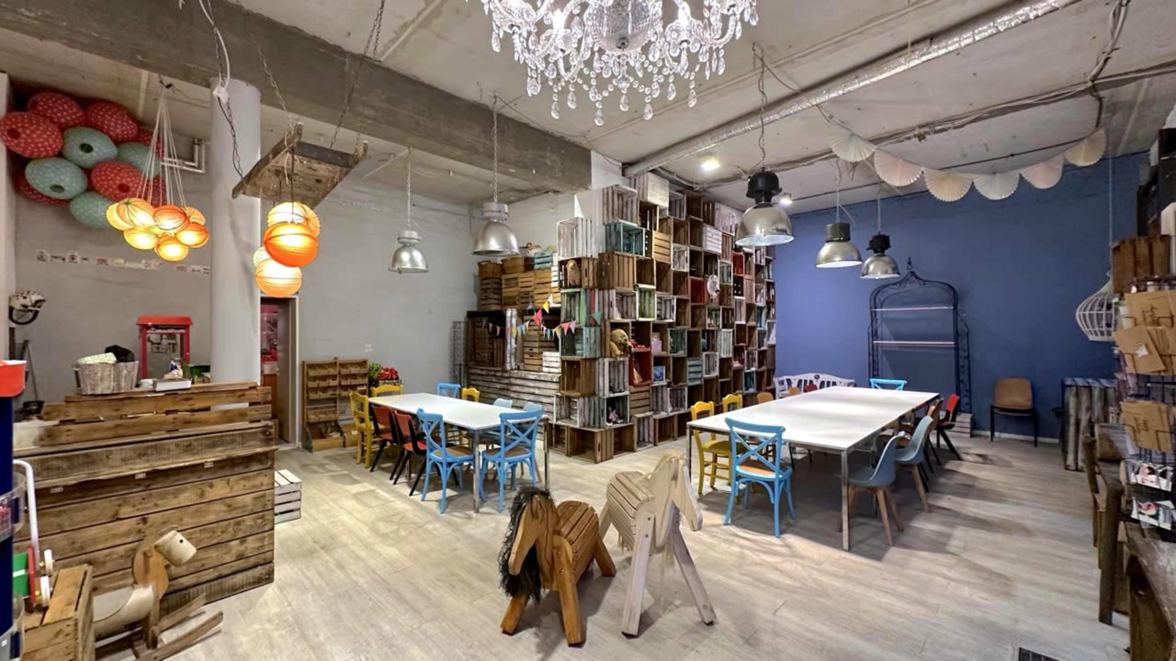 The image shows a colorful children's activity space featuring wooden furniture, shelves filled with crates, and a chandelier hanging from the ceiling.