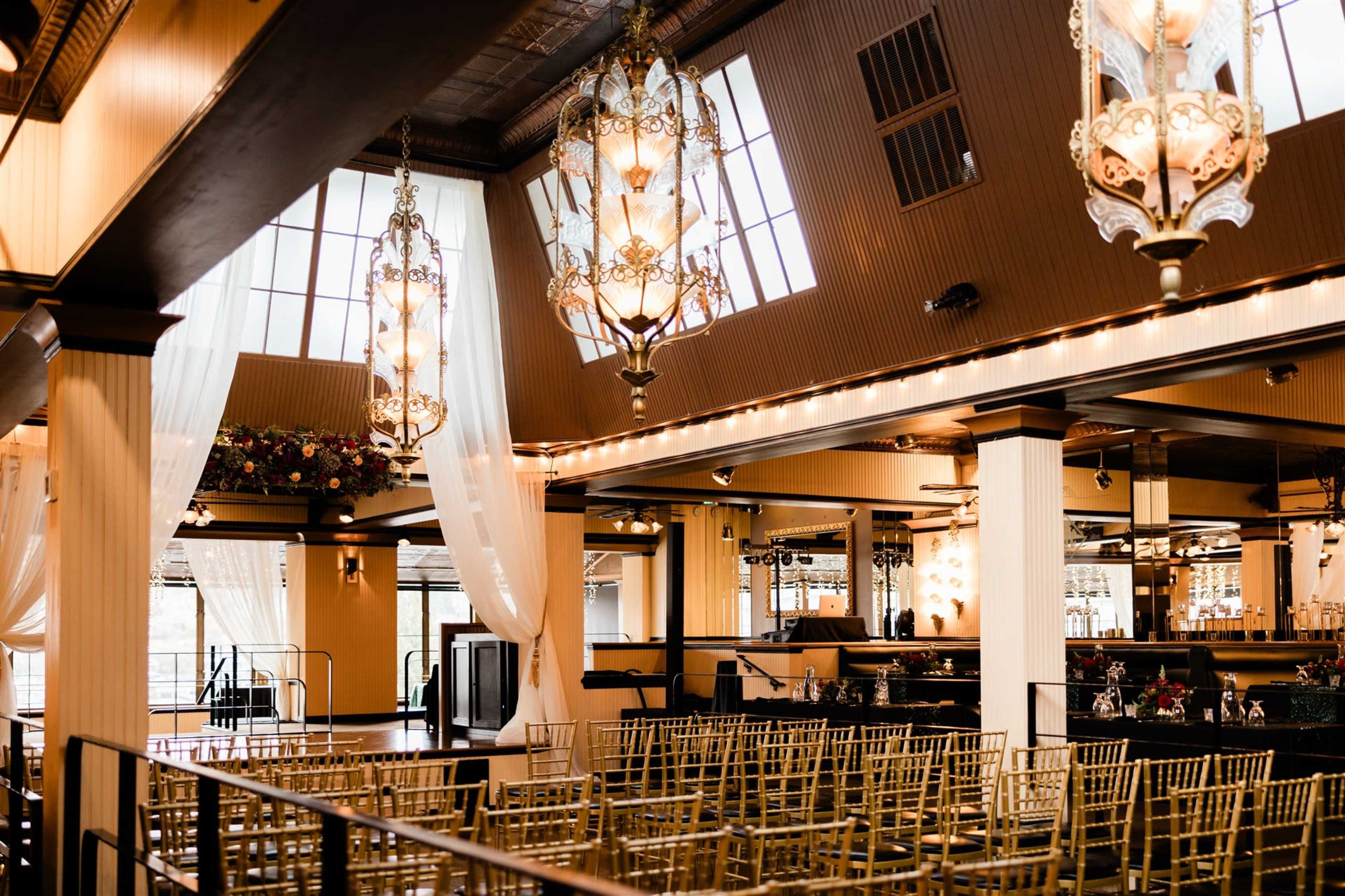The image shows a wedding venue with gold chiavari chairs arranged in rows, elegant chandeliers hanging from the ceiling, and large windows allowing natural light to enter.