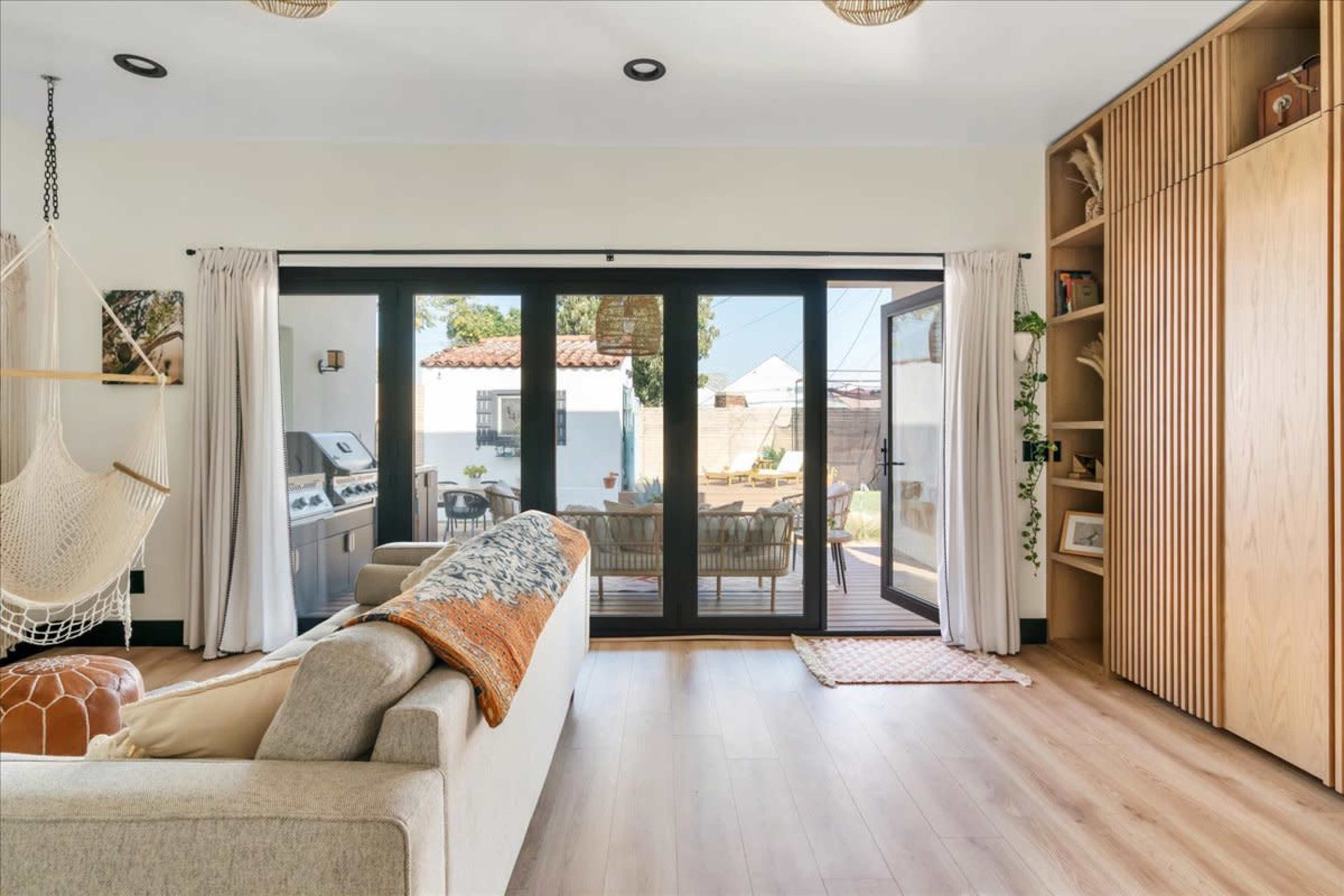 The image shows a modern living room with a beige sofa, a hammock, and large glass doors leading to an outdoor patio area.