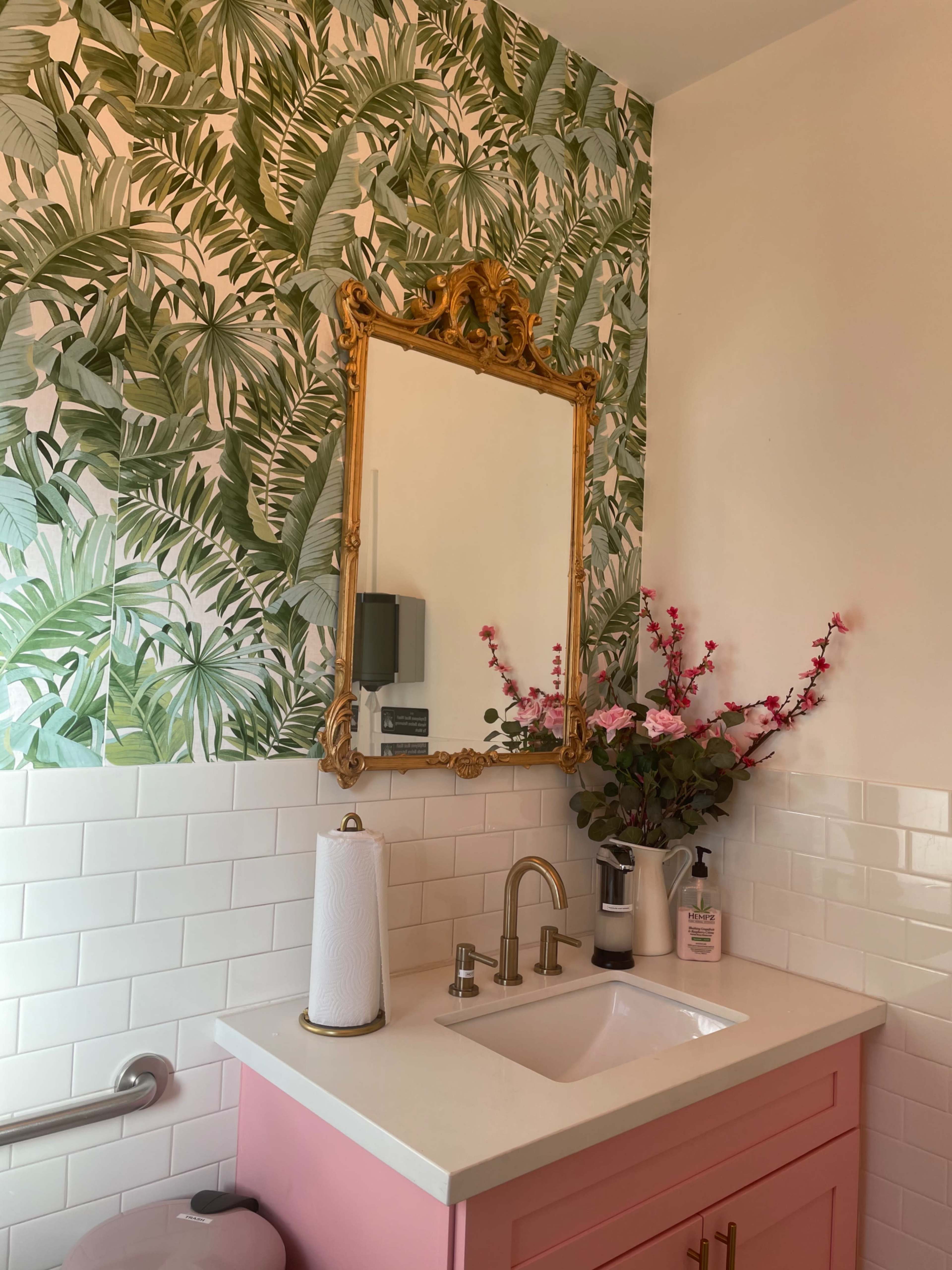 A bathroom features a pink vanity with a white sink, a gold-framed mirror, and a wall adorned with green leaf-patterned wallpaper.
