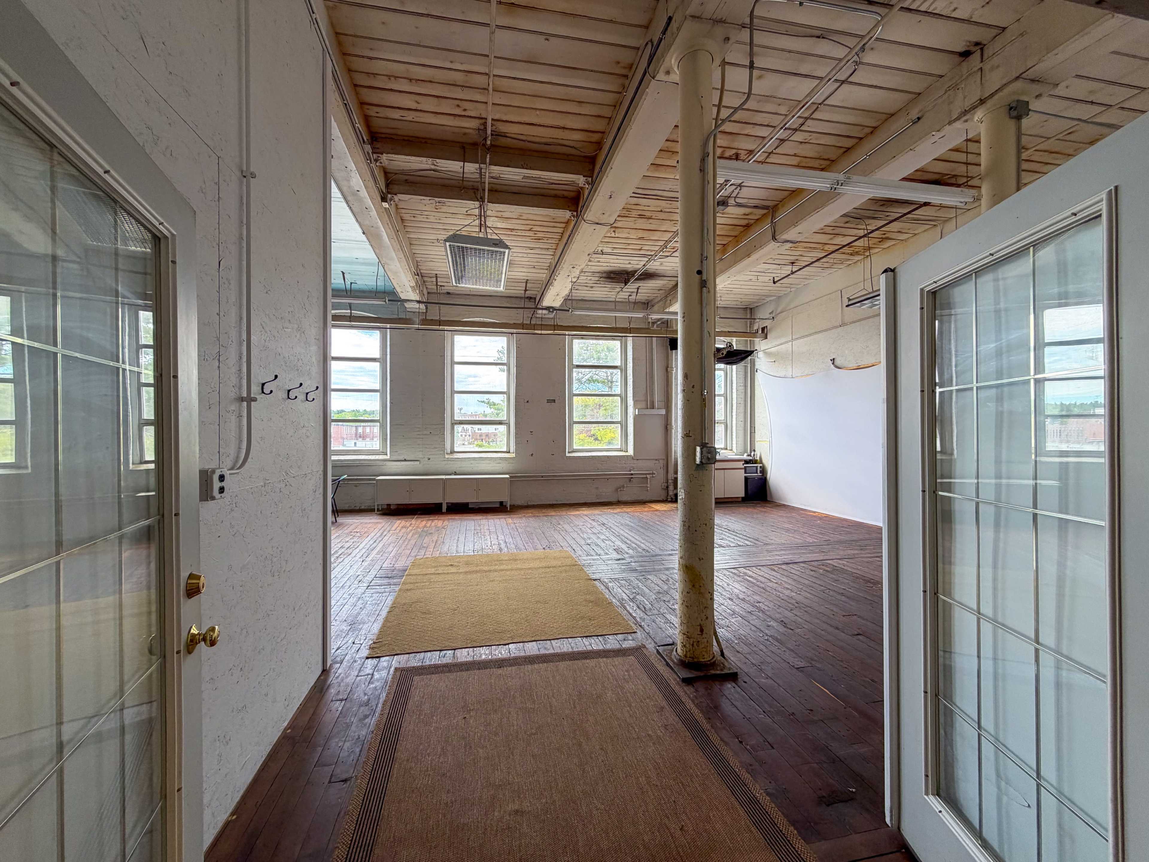The image shows a spacious, industrial-style room with large windows, exposed wooden beams, and hardwood flooring, featuring a clear entryway leading into the space.