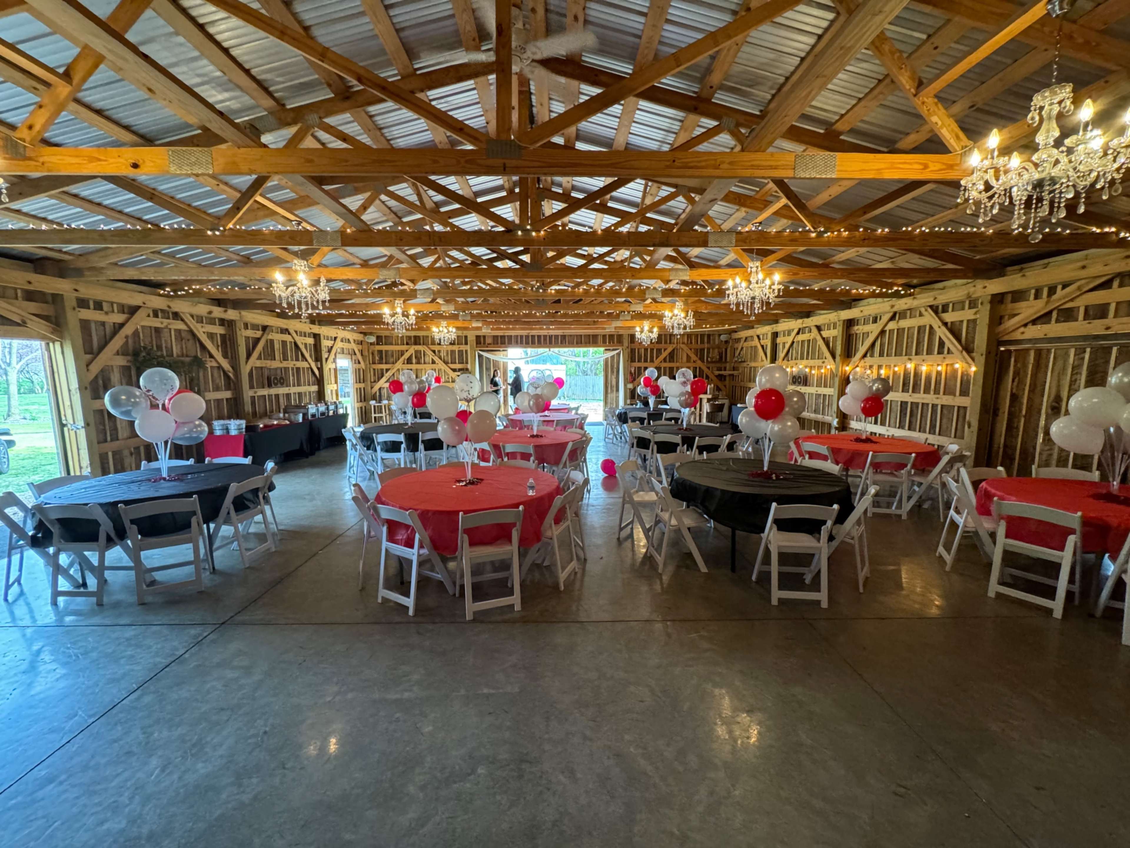 The interior of a barn is decorated for an event, featuring round tables with red and black tablecloths, and balloons in red and white.
