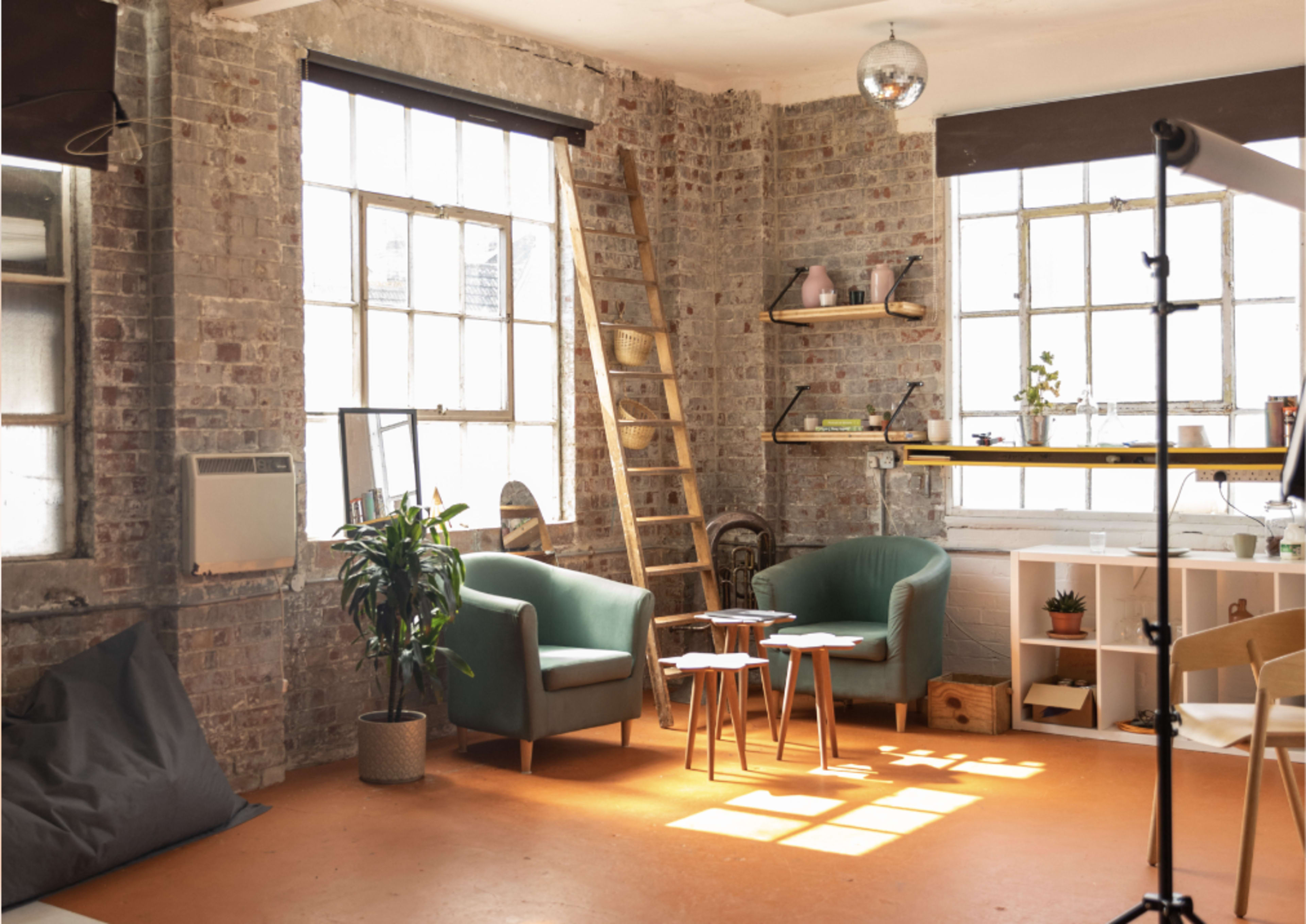 The scene shows a cozy room with brick walls, two green armchairs, a small wooden ladder, and shelves containing decorative items, illuminated by natural light from large windows.