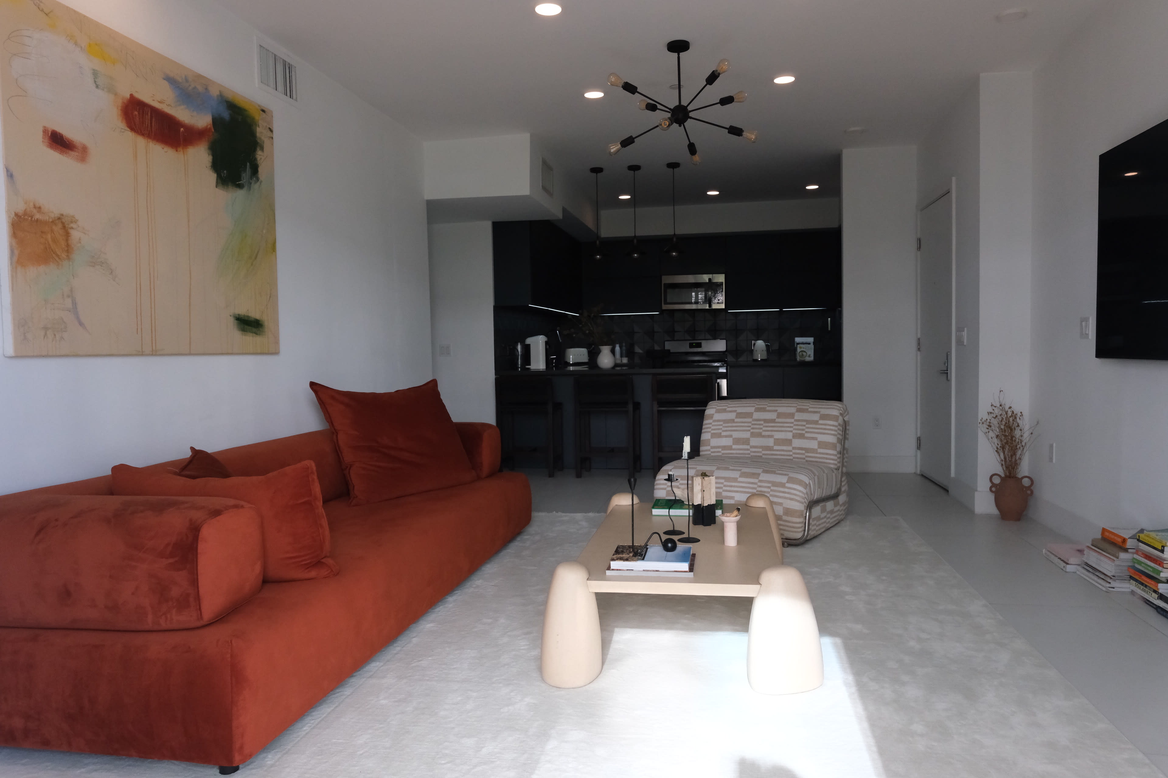 The image depicts a modern living room featuring a large brown sofa, a light-colored rug, a minimalist coffee table, and a dining area in the background with black cabinetry.