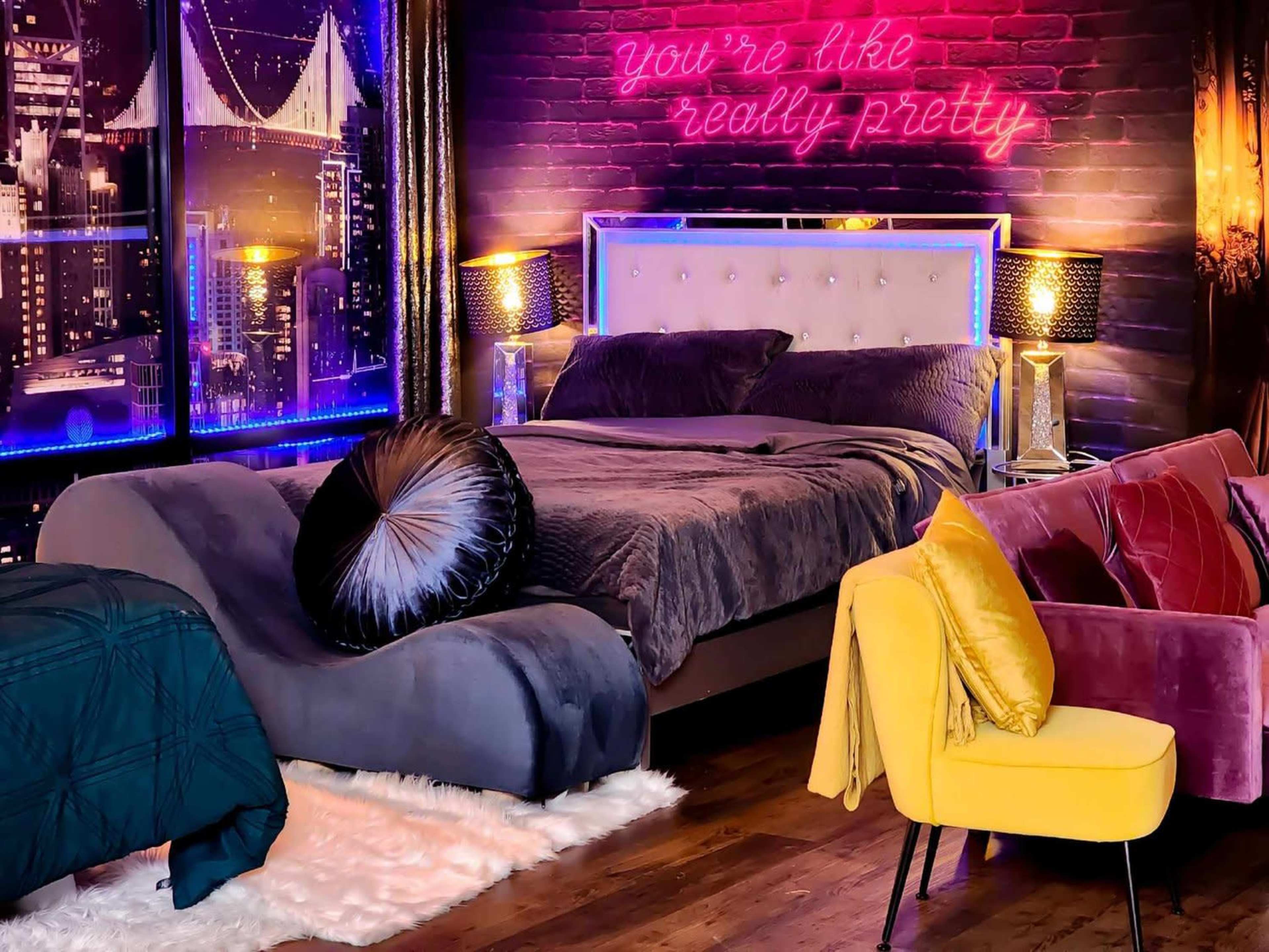 A stylish bedroom with a bed, a plush lounge chair, and a neon sign on the wall that reads "you're like really pretty."