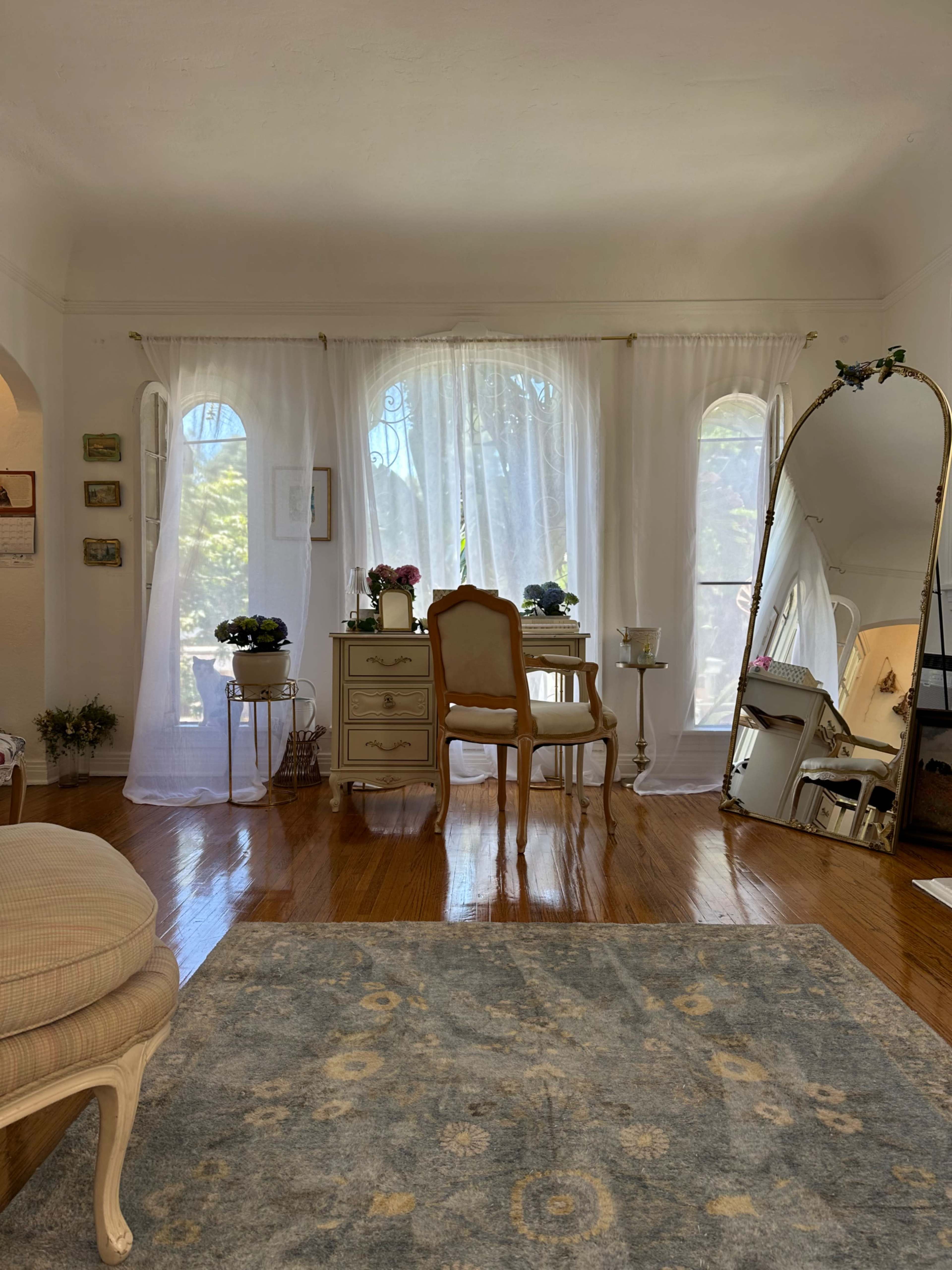 A bright room features a small table and chair near large windows with sheer curtains, a decorative mirror, and a rug on the hardwood floor.