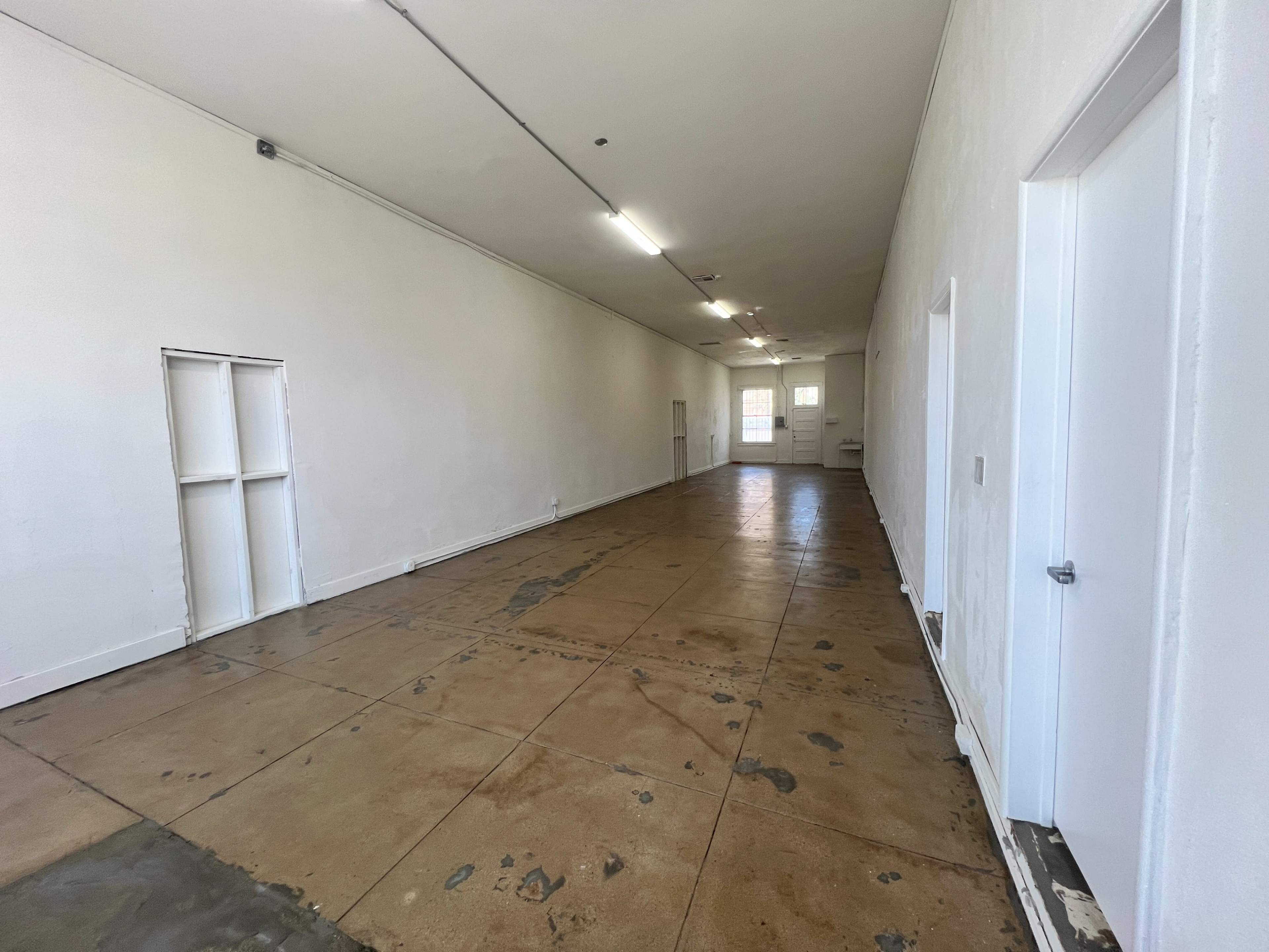 An empty room with a long, bare floor and white walls, featuring a door on one side and a small opening in the wall.