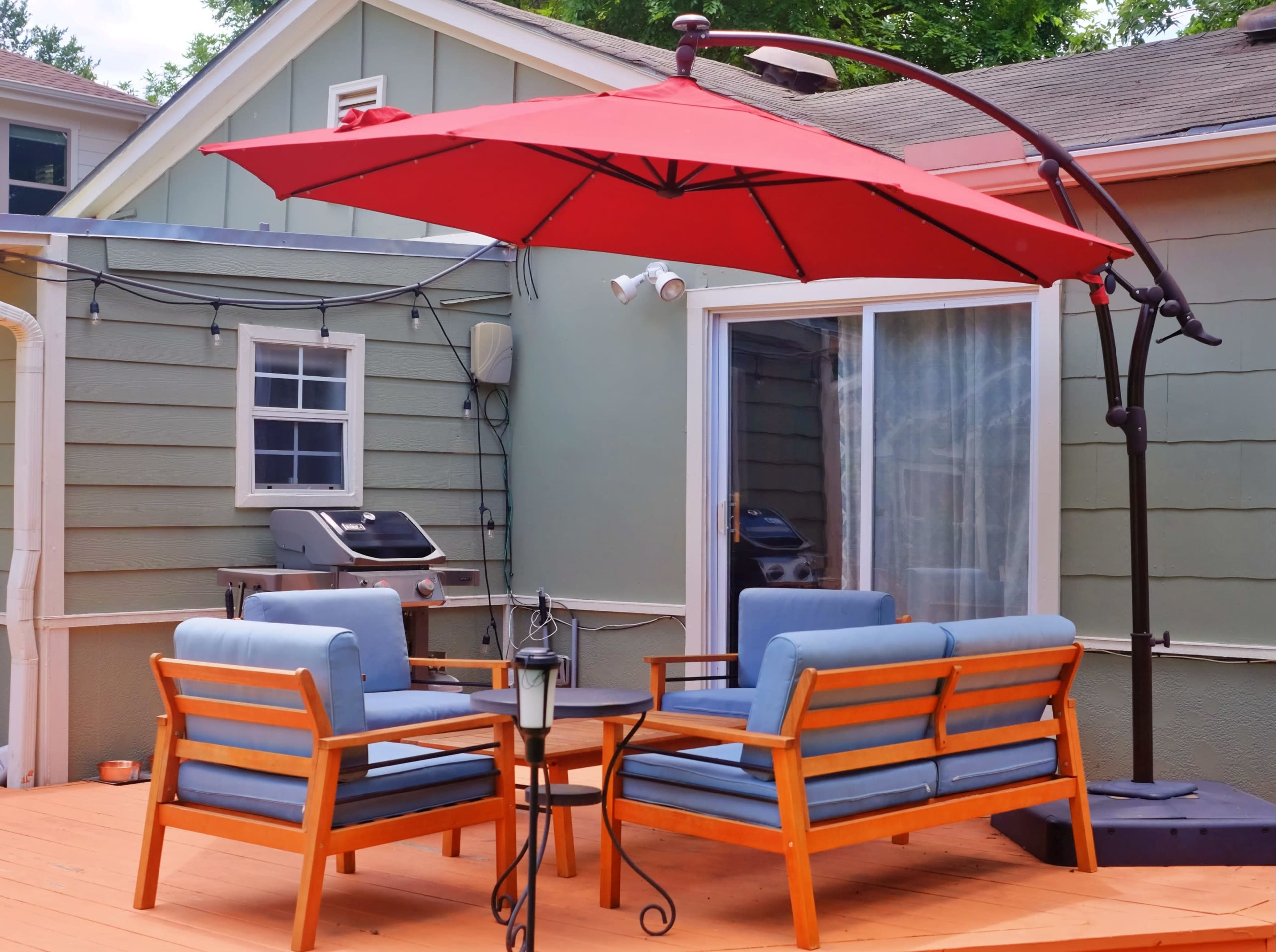 The image shows a patio area featuring two blue cushioned chairs, a small table, and a large red umbrella, with a grill and a window visible in the background.