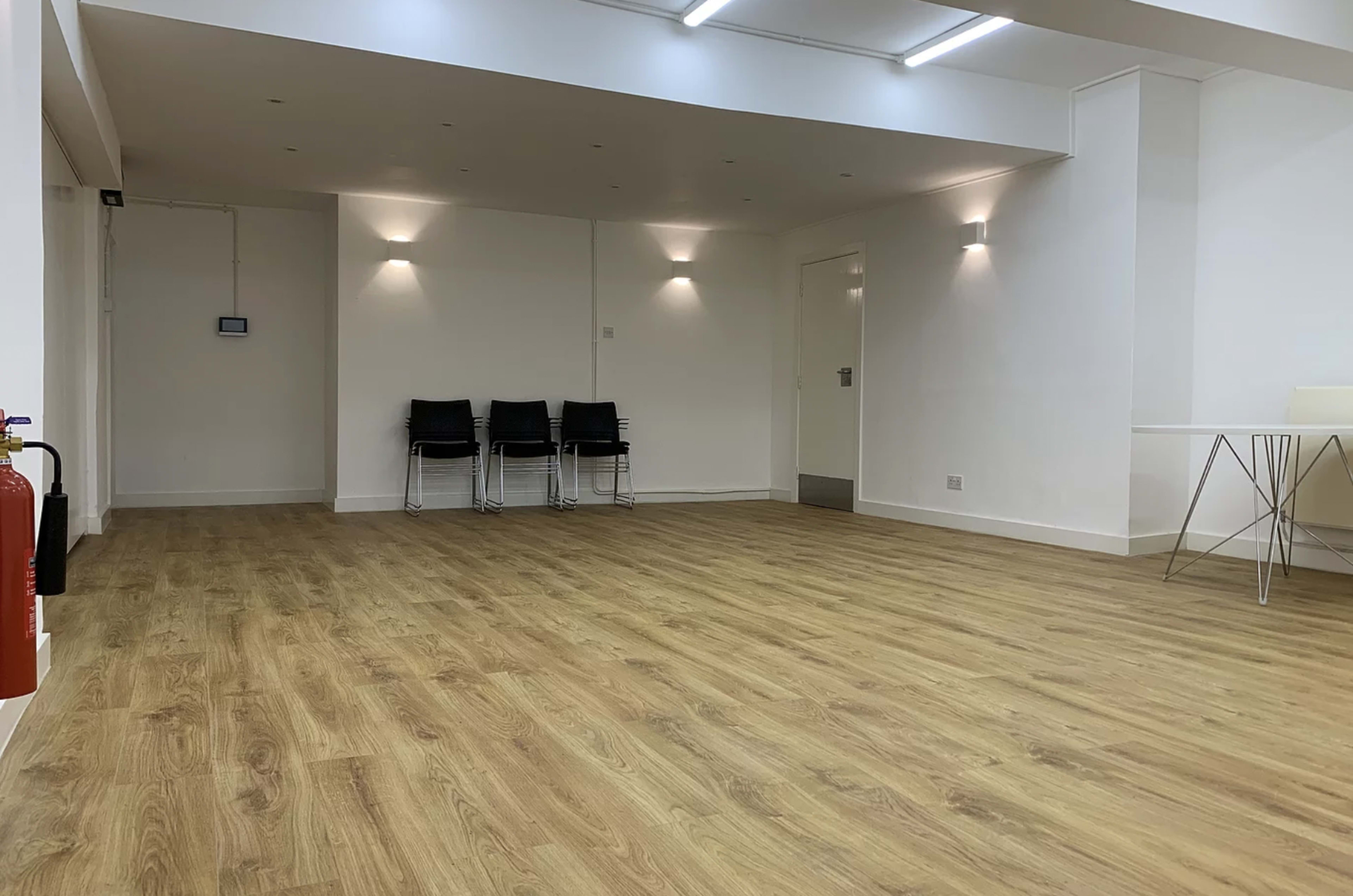 The image shows a spacious, empty room featuring wooden flooring, white walls, and a few black chairs lined against one wall.