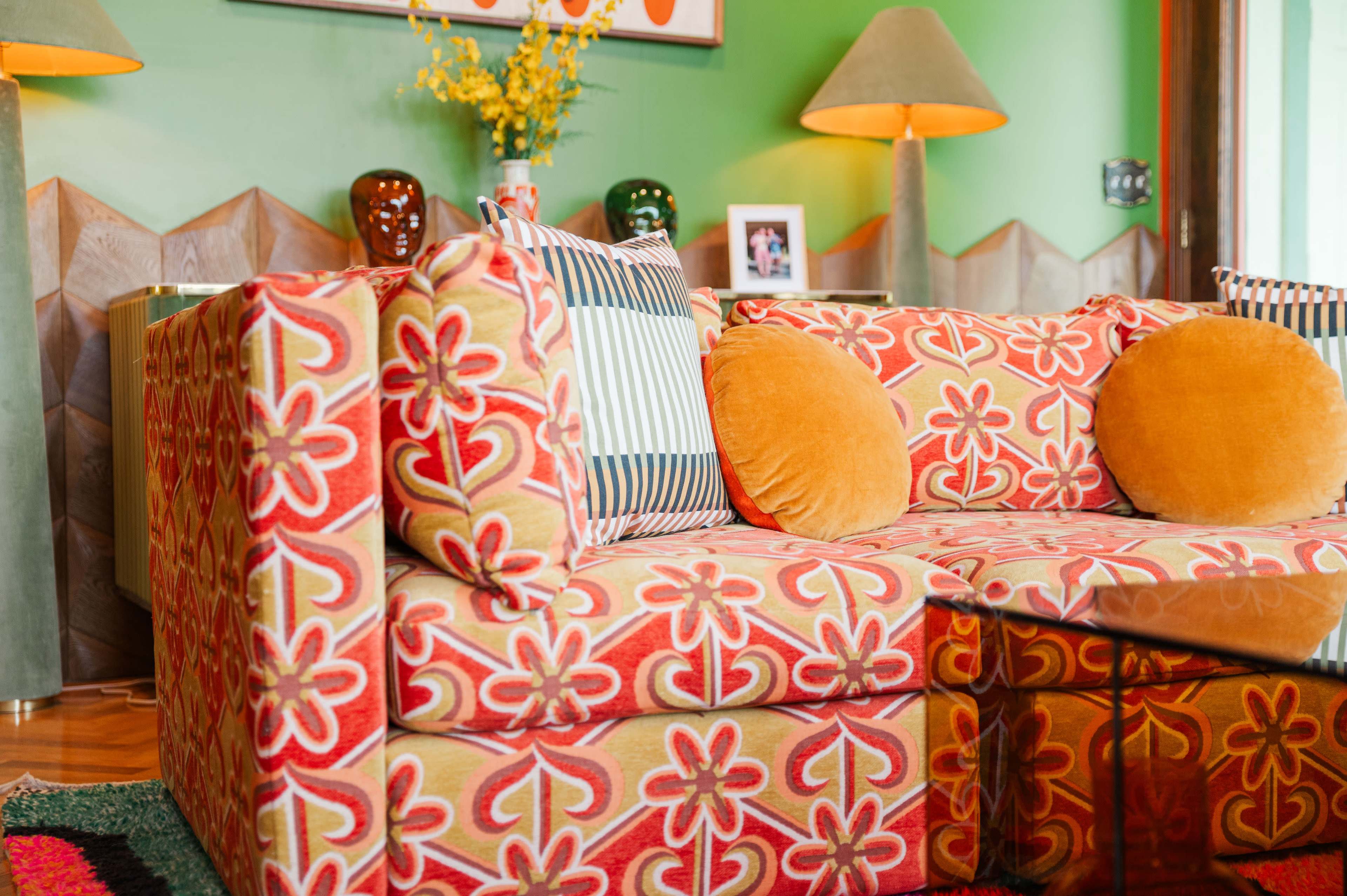The image shows a colorful couch with floral-patterned upholstery and orange cushions, set in a room with green walls and decorative objects.