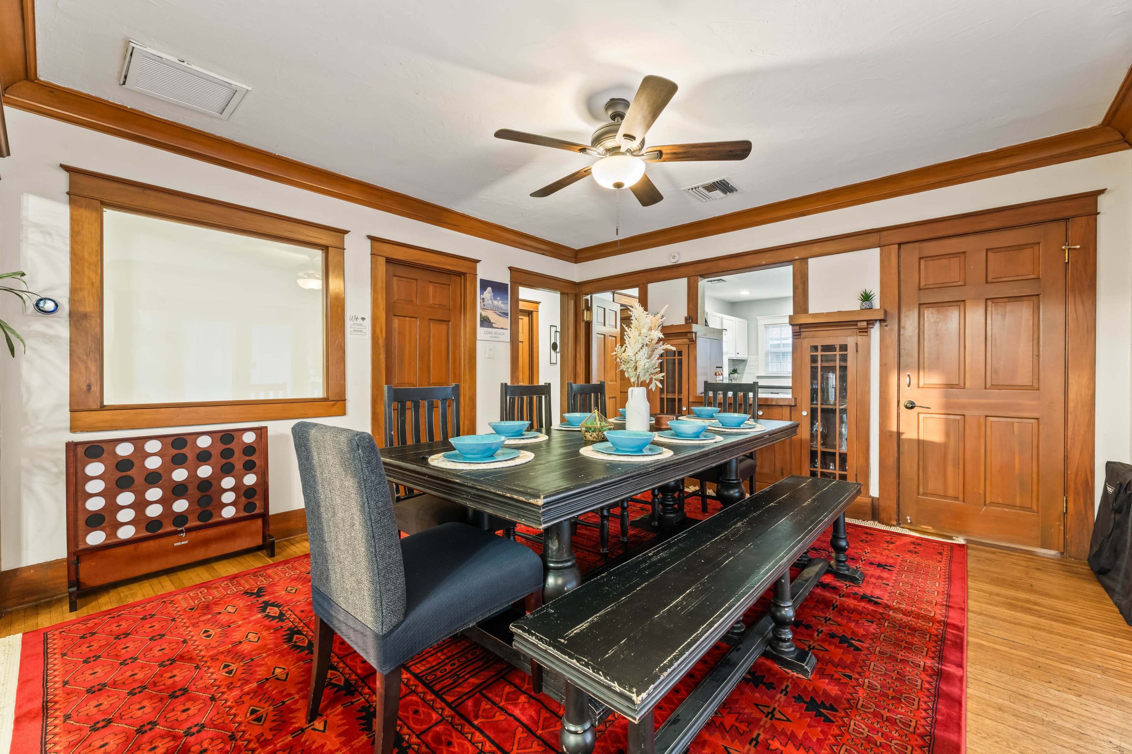 The image shows a dining room featuring a large black wooden table surrounded by dark wooden chairs and a bench, with a fan overhead and a red patterned rug on the floor.