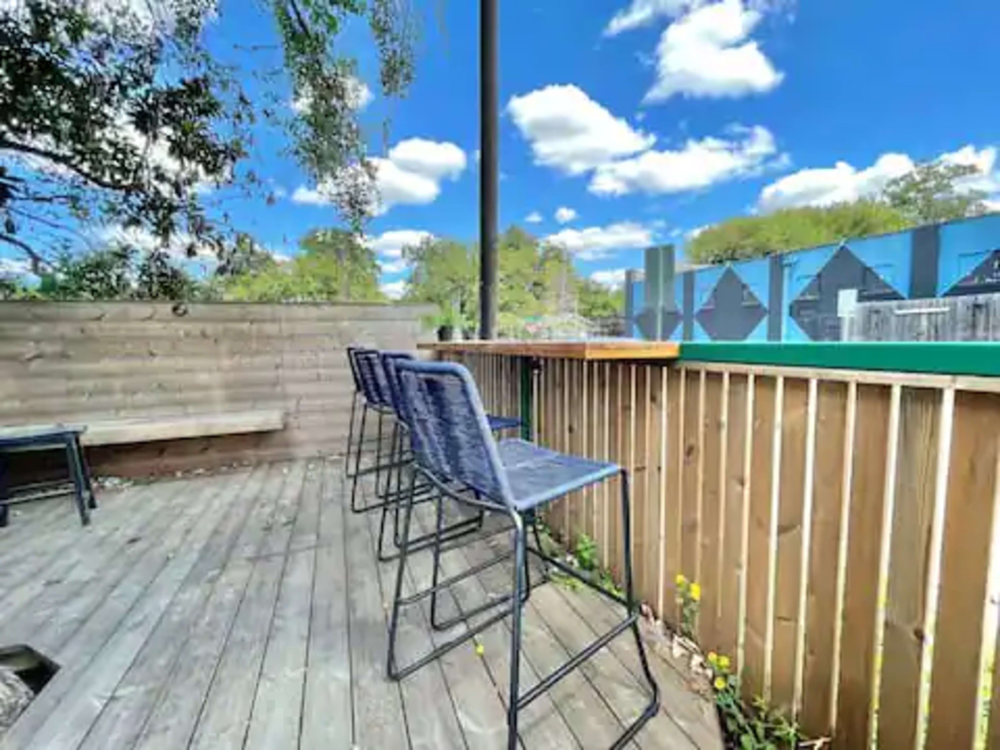 A wooden deck with three high stools facing a green area under a blue sky with clouds.