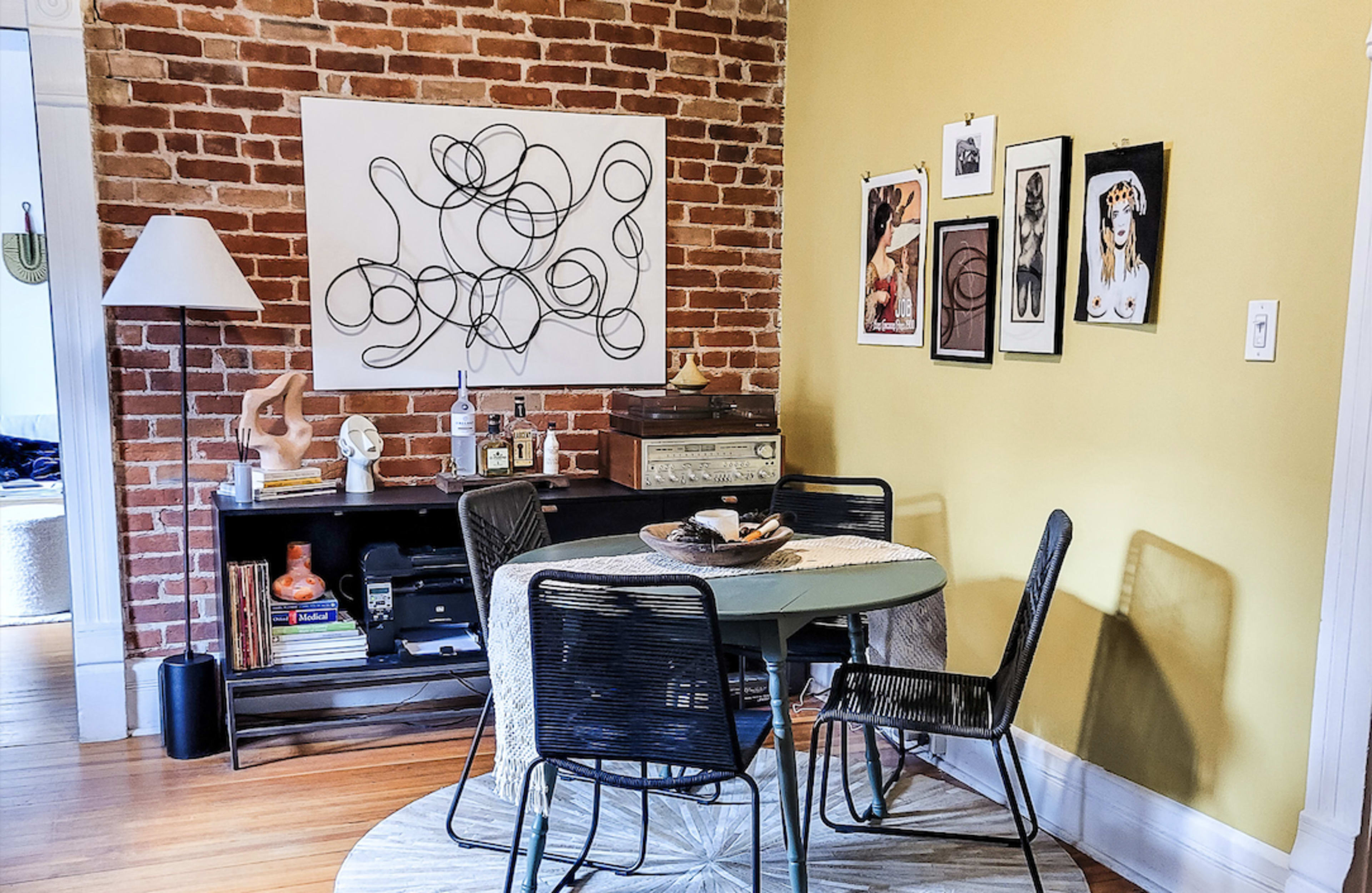The image shows a dining area with a round table and four black chairs against a yellow wall, featuring framed artwork and a brick accent wall.