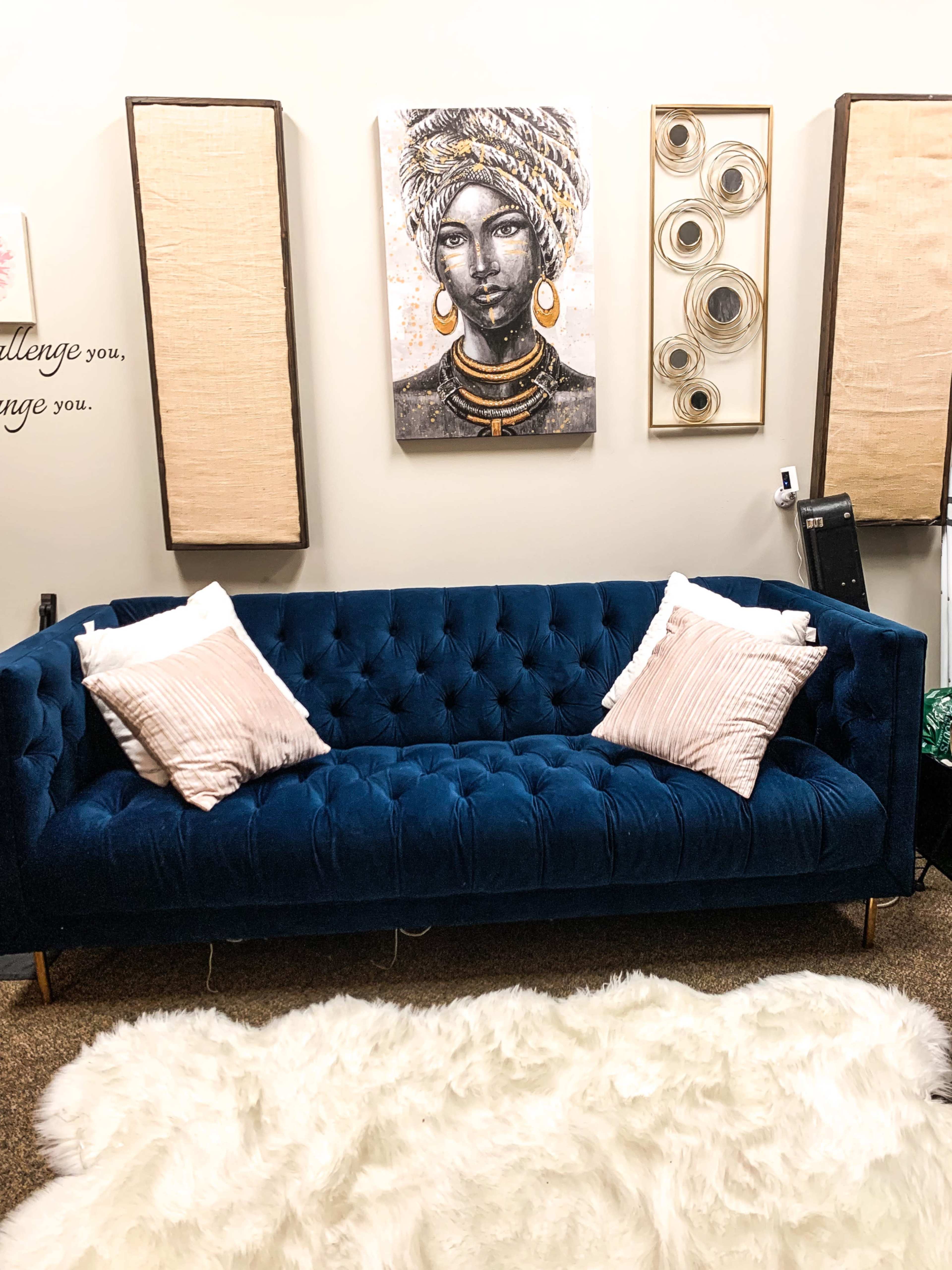 The image features a blue tufted sofa with two light-colored decorative pillows, positioned on a textured beige rug beneath a wall adorned with artistic decorations.