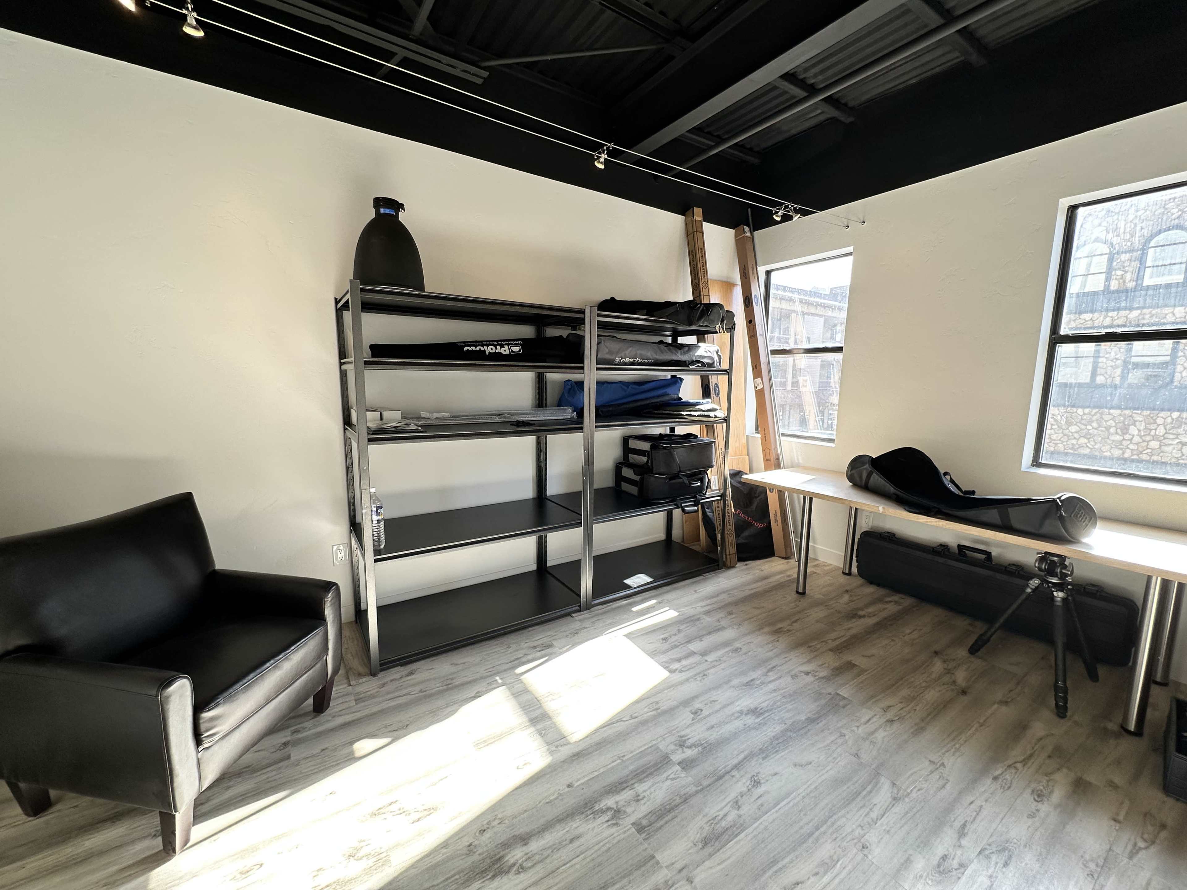 A sparsely furnished room with a black chair, a shelving unit filled with equipment, and a large table beside a window.
