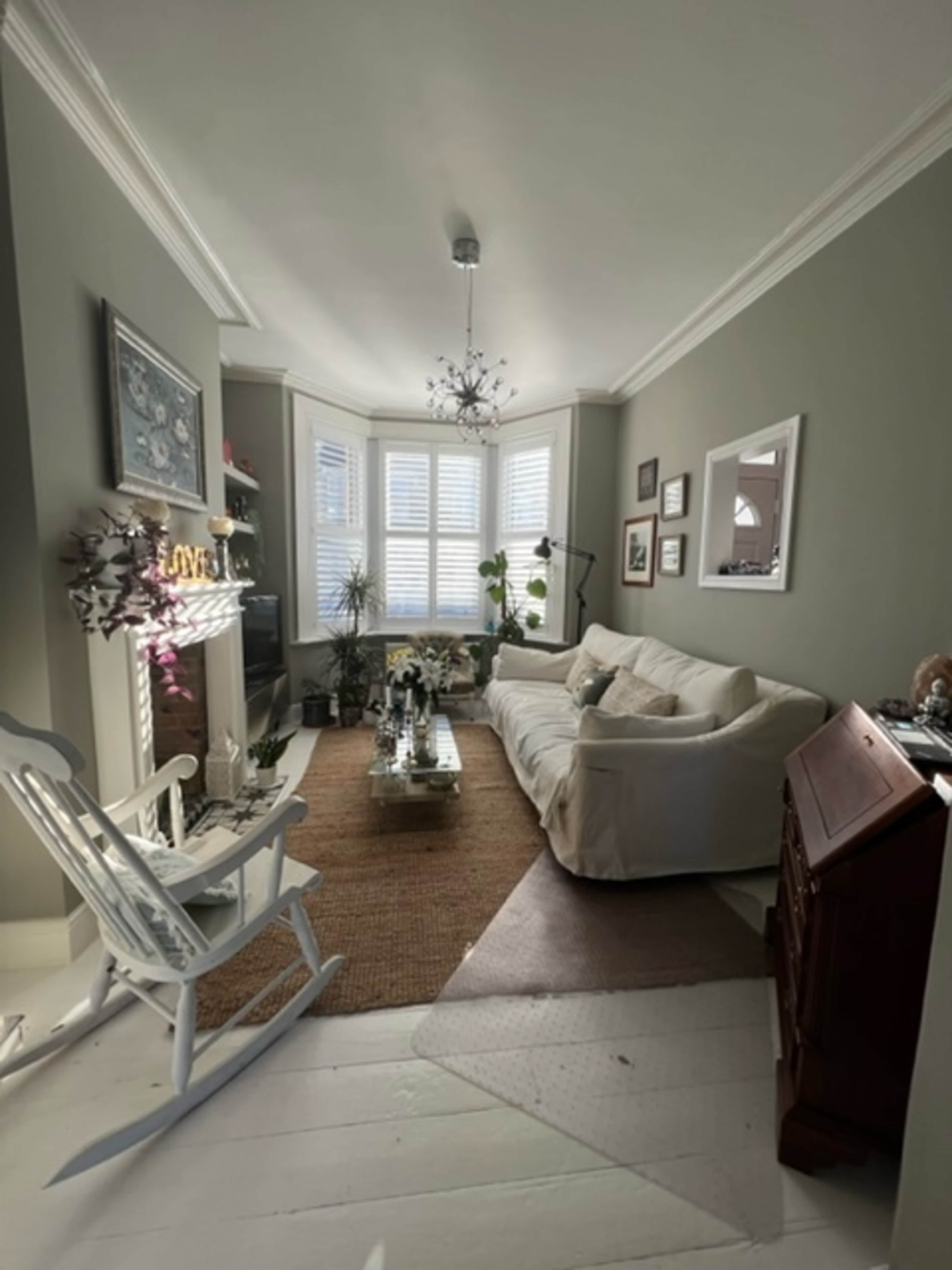 A living room featuring a light-colored sofa, a glass coffee table, a rocking chair, and a decorative fireplace, with natural light coming through bay windows.