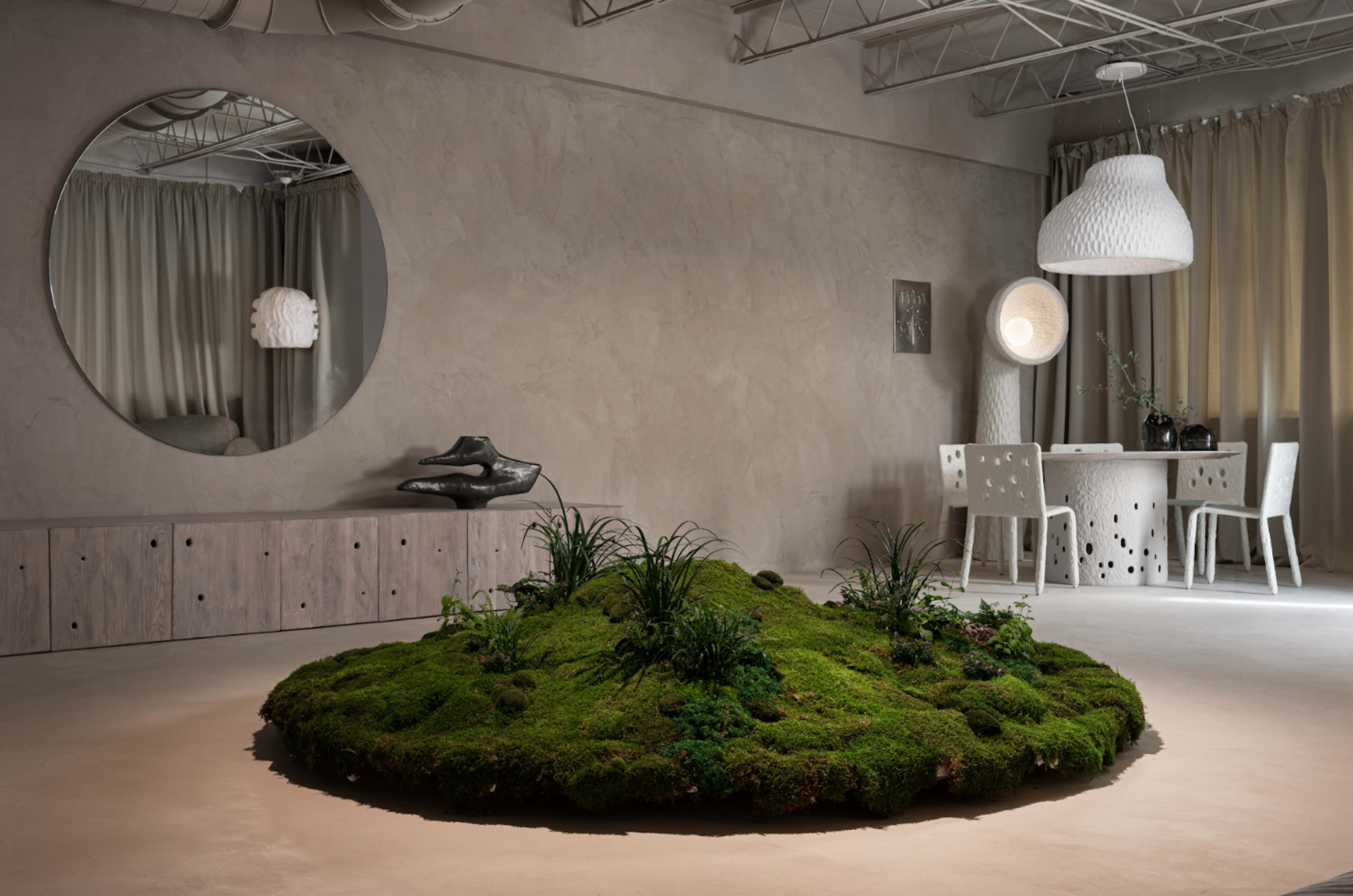 The image shows a minimalist interior space featuring a large circular patch of moss in the center, surrounded by modern furniture and decorative elements.