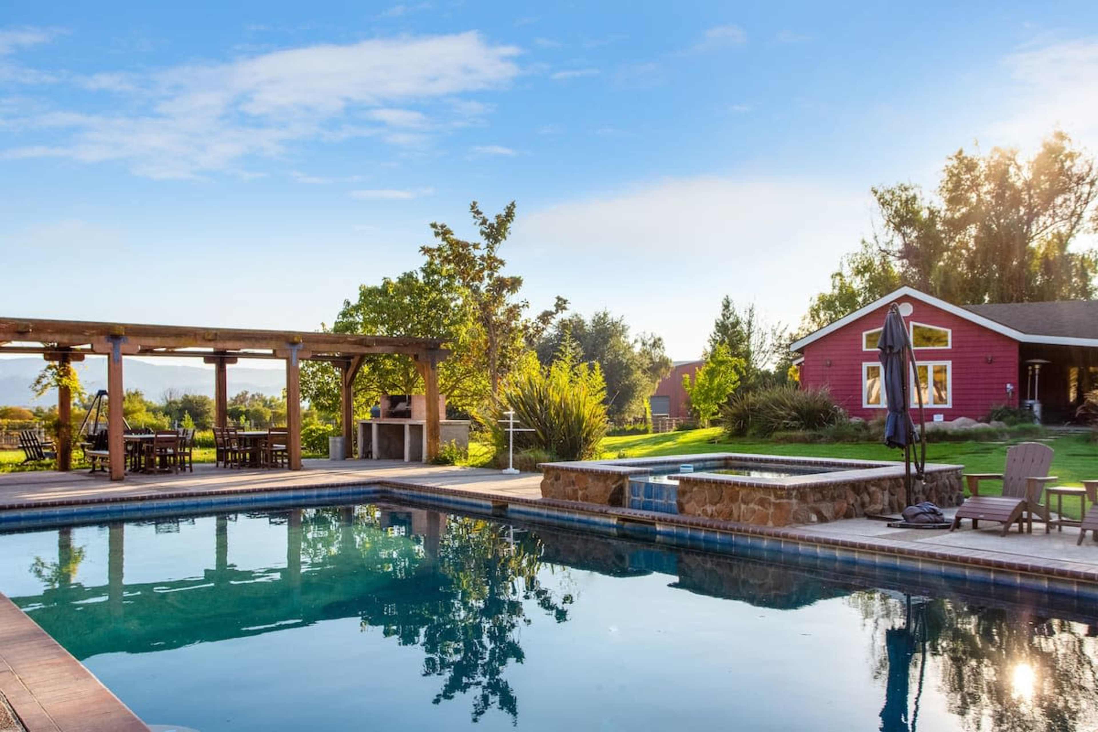 A landscaped backyard features a swimming pool, a wooden pergola with seating, and a red house surrounded by trees and grass.