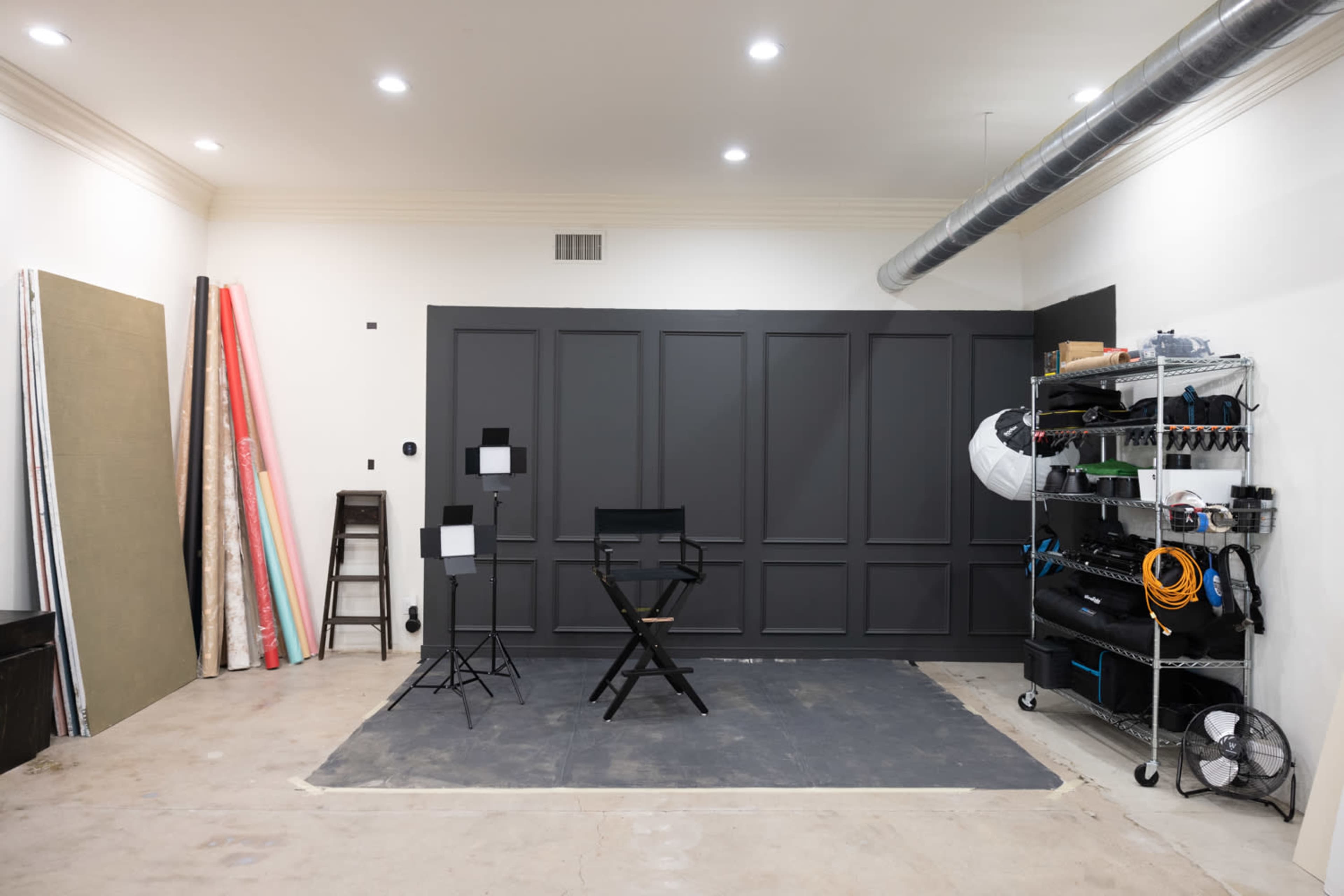 A well-organized photography studio features a gray wall, a director's chair on a black mat, lighting equipment, and a shelving unit with various tools and materials.