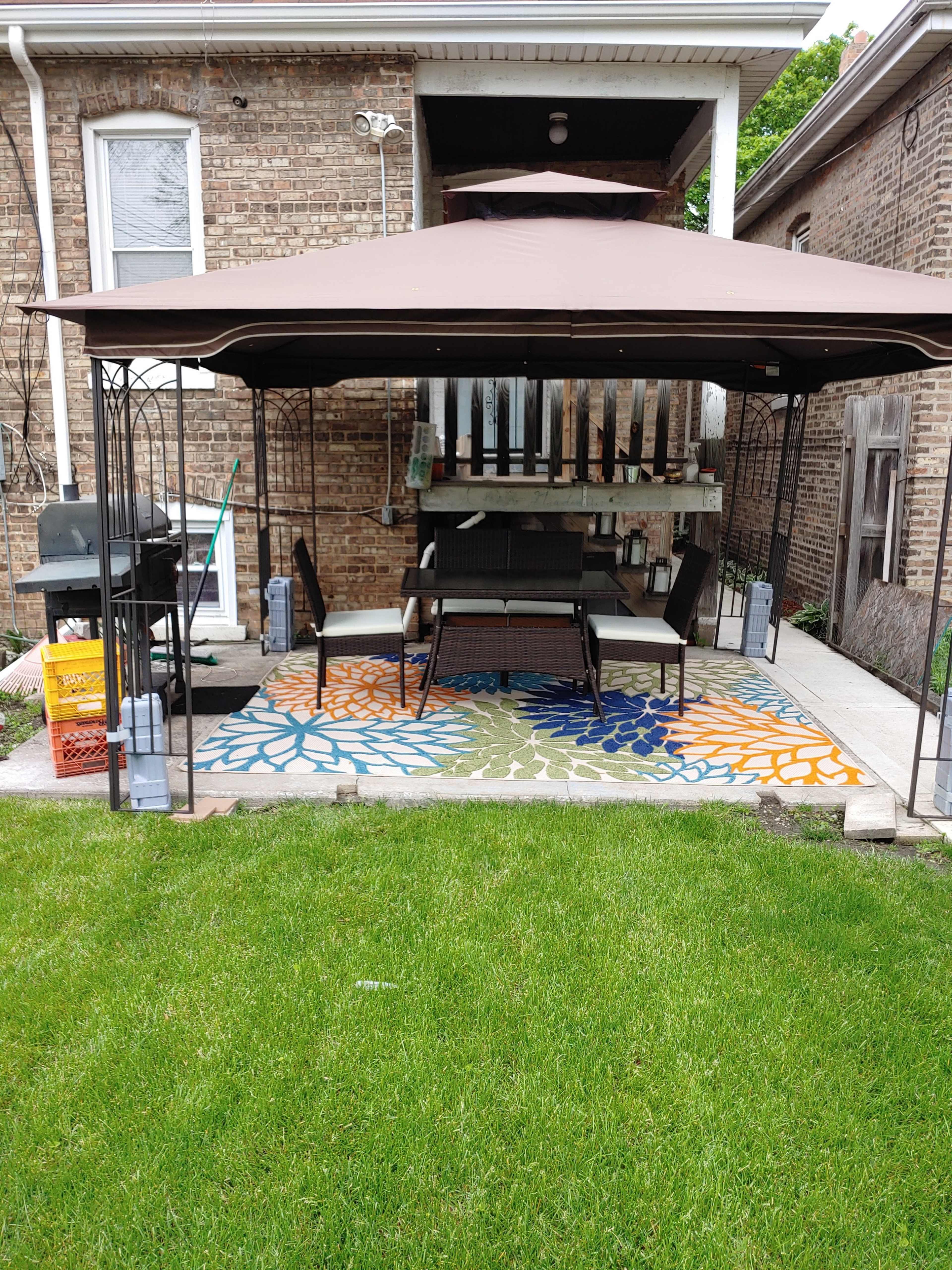 A metal gazebo with a brown canopy shelters a seating area on a patterned rug in a backyard with grass and nearby cooking equipment.
