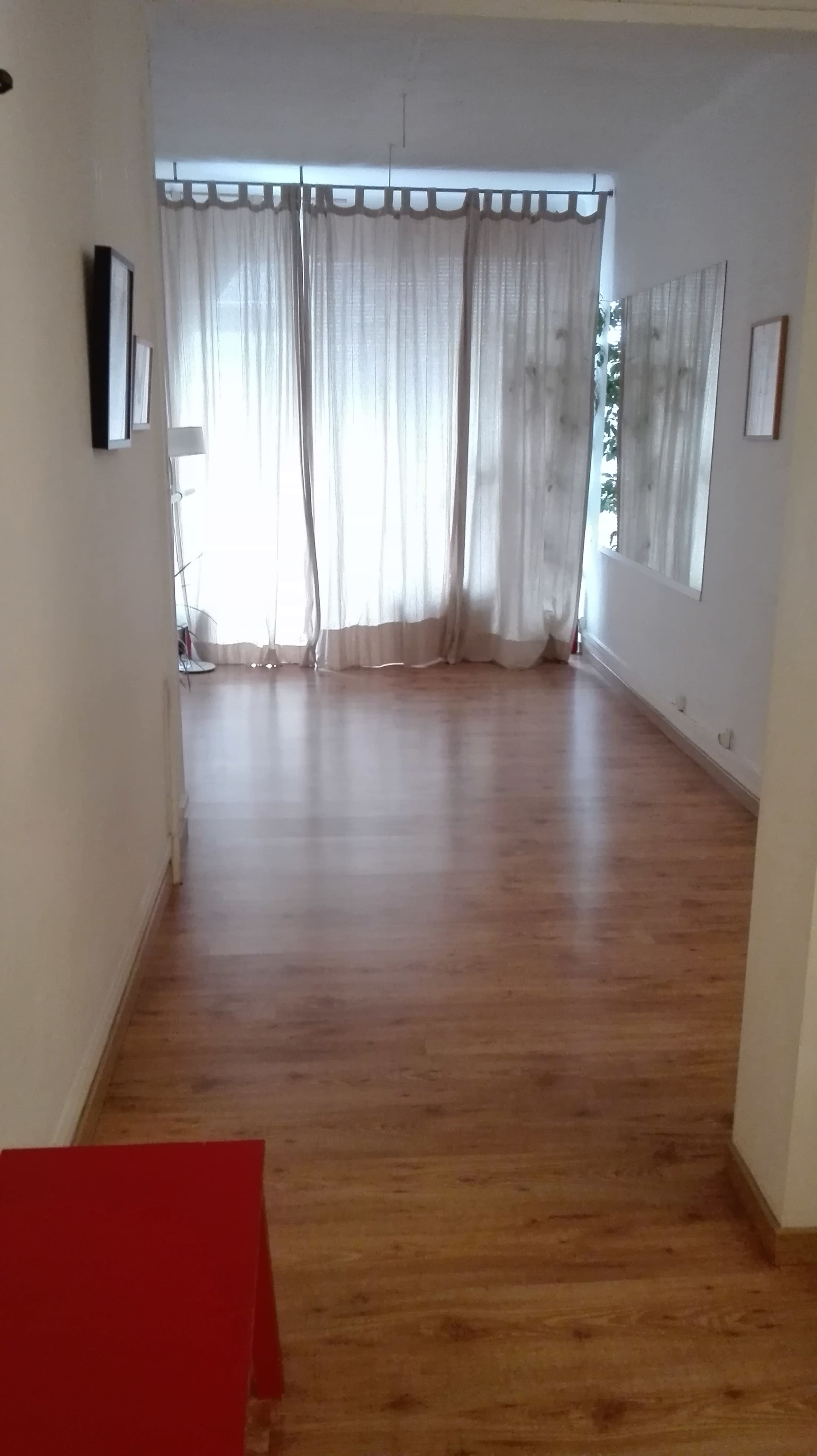 A narrow hallway leads to a bright room with sheer curtains and wooden flooring.