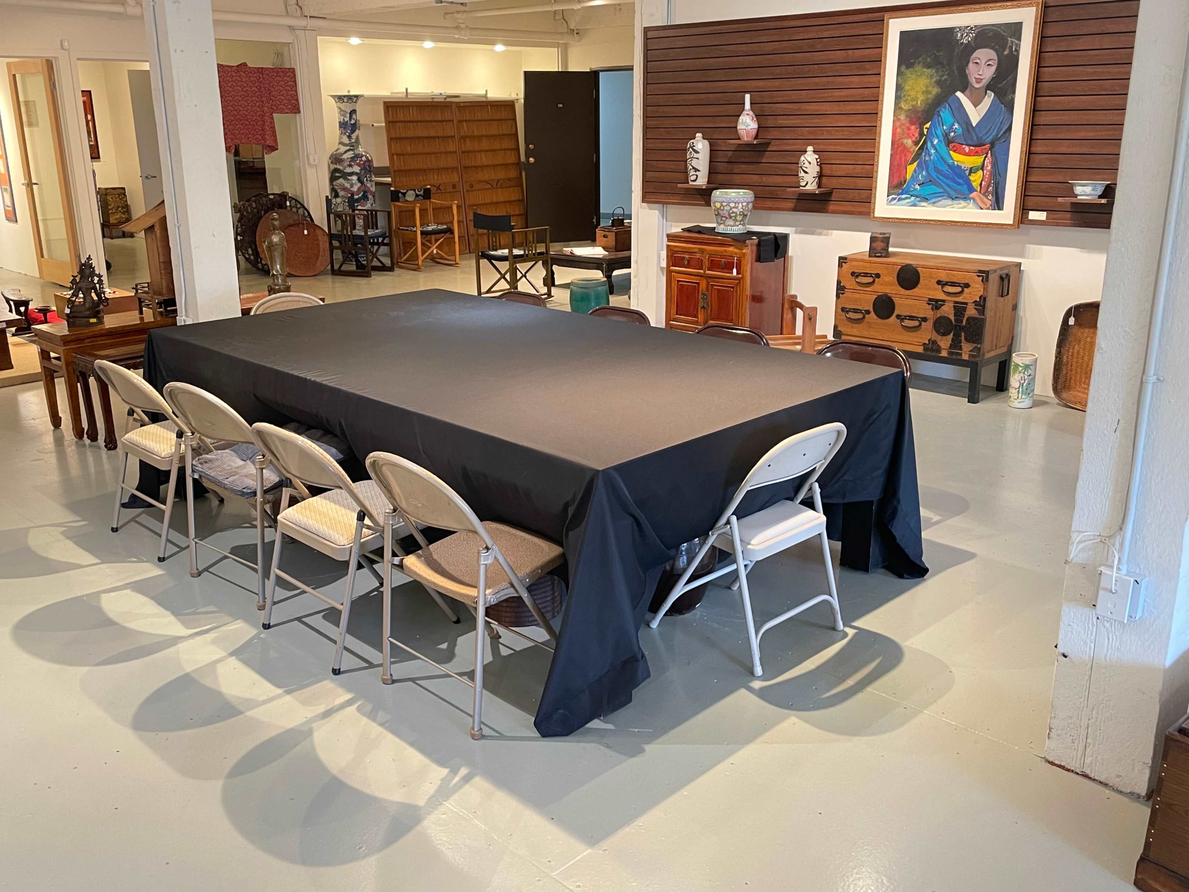 A large rectangular table with a black tablecloth is surrounded by plastic chairs in a spacious indoor area filled with various decorative items and furniture.
