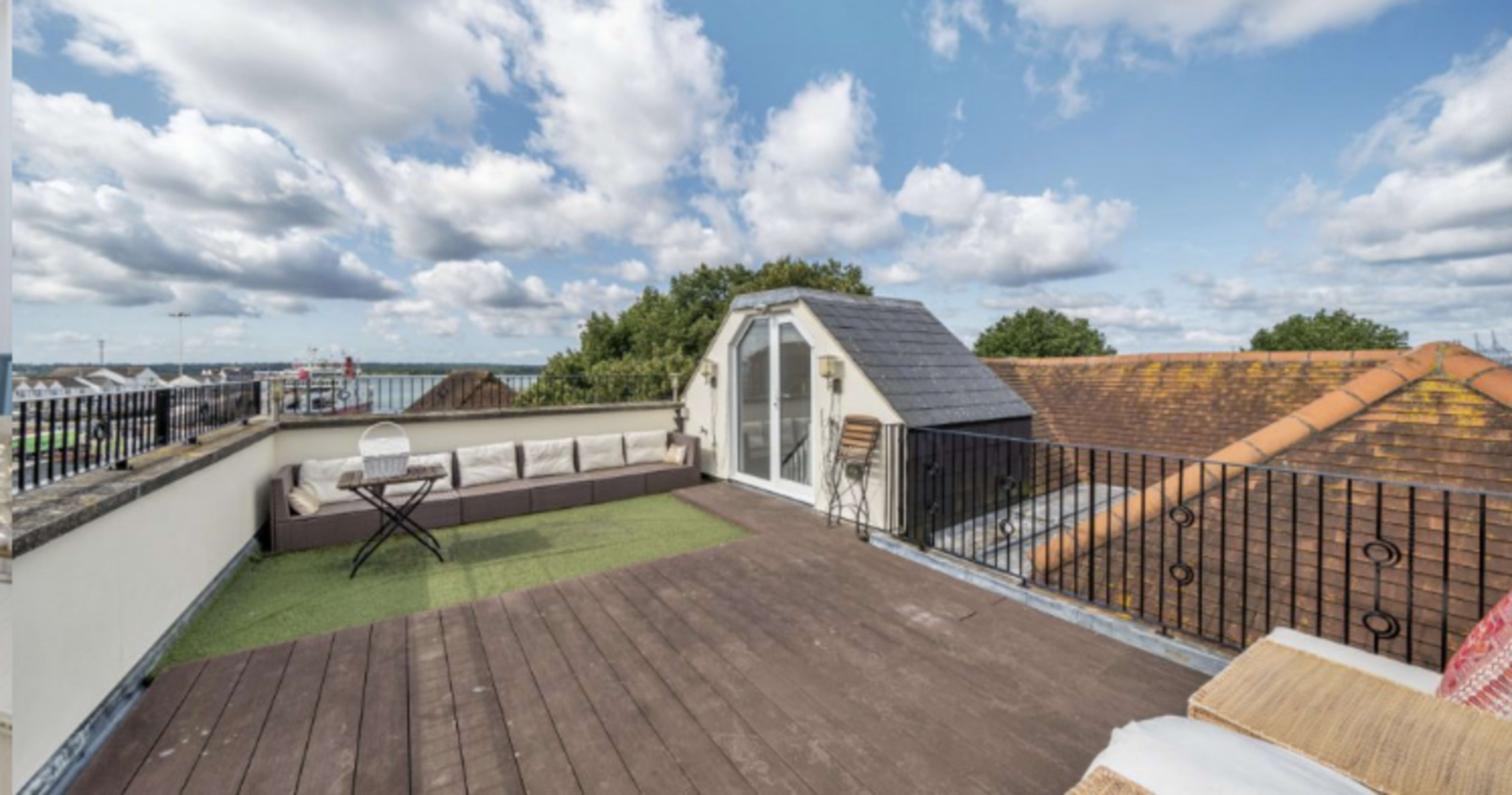 A rooftop terrace with a wooden floor, green grass, and a white structure, surrounded by a railing and overlooking a cloudy sky.