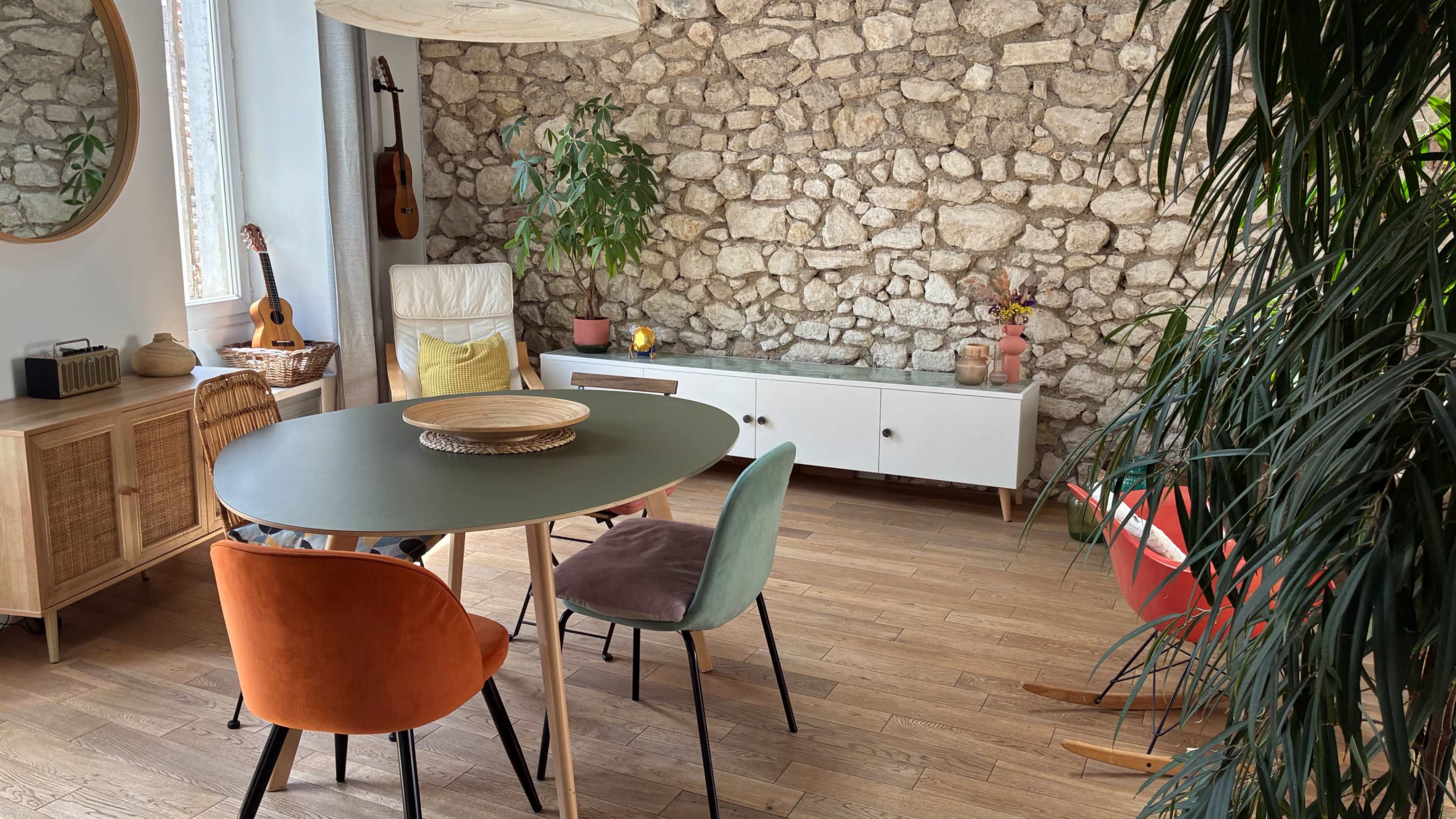 The image shows a modern dining area with a circular table surrounded by colorful chairs, a stone wall, a potted plant, and various decorative elements.