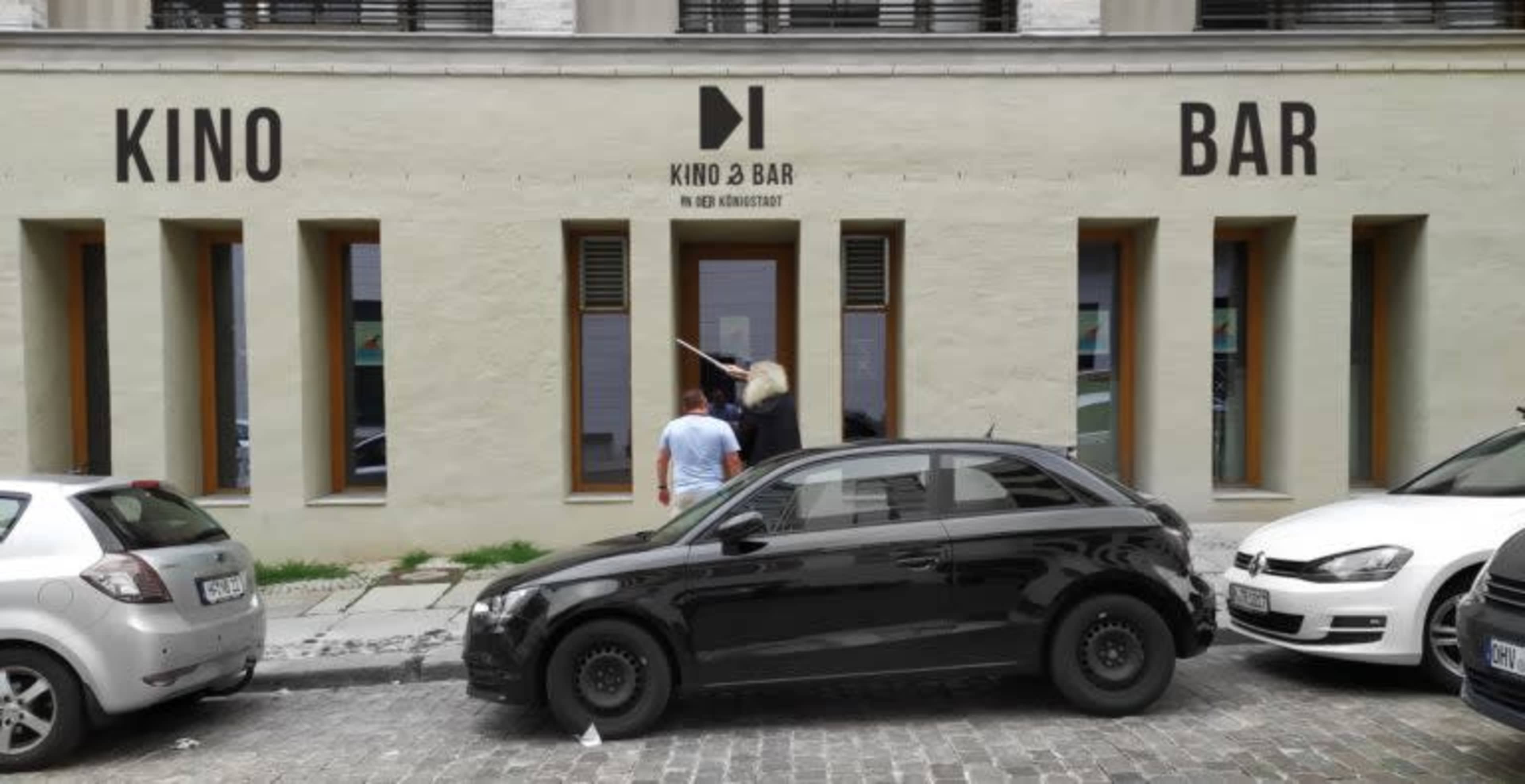 A couple stands outside a building with the words "KINO" and "BAR" prominently displayed above the entrance.