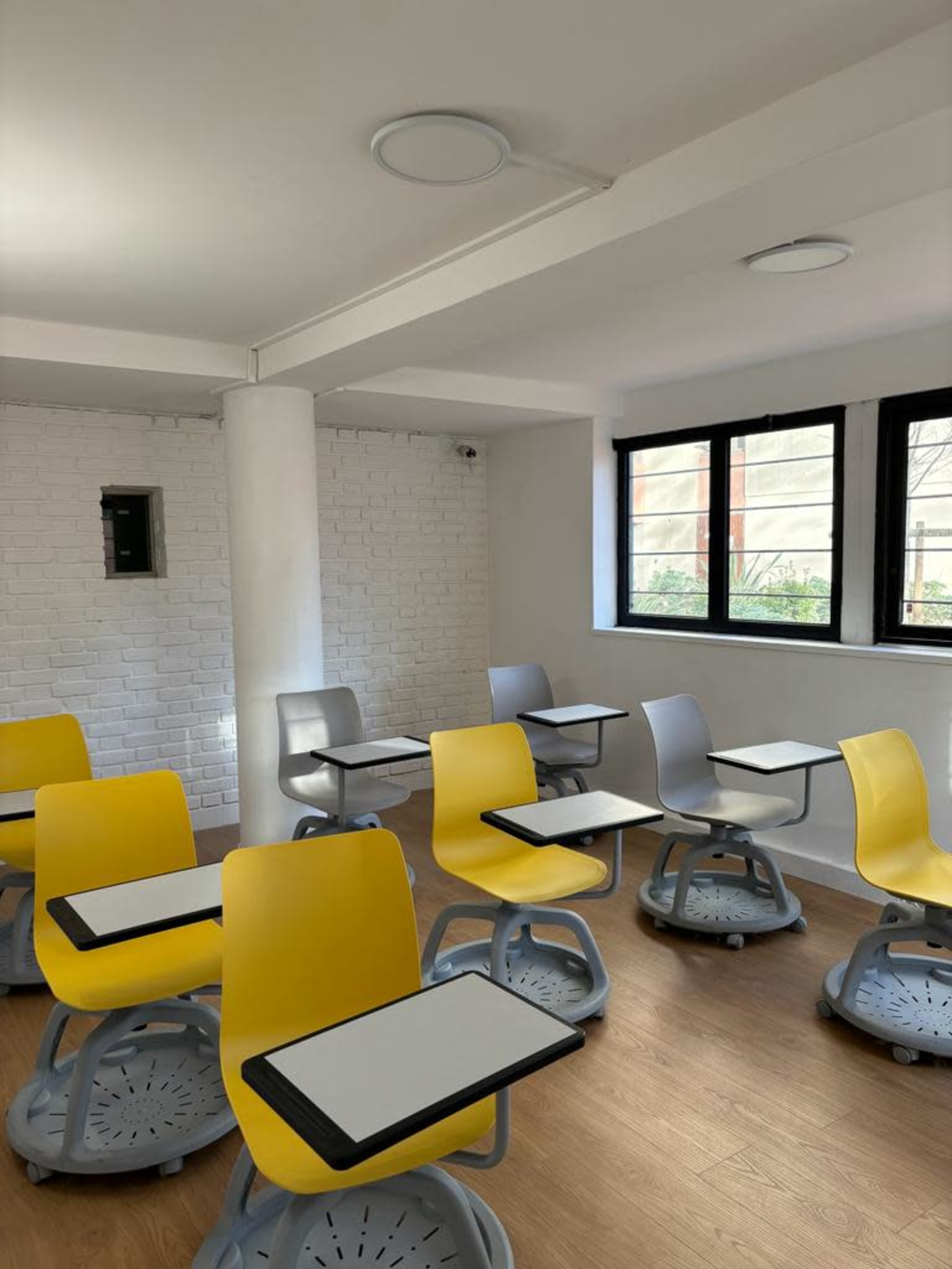 A modern classroom with yellow and gray chairs arranged around small tables, set against a bright, minimalist interior.