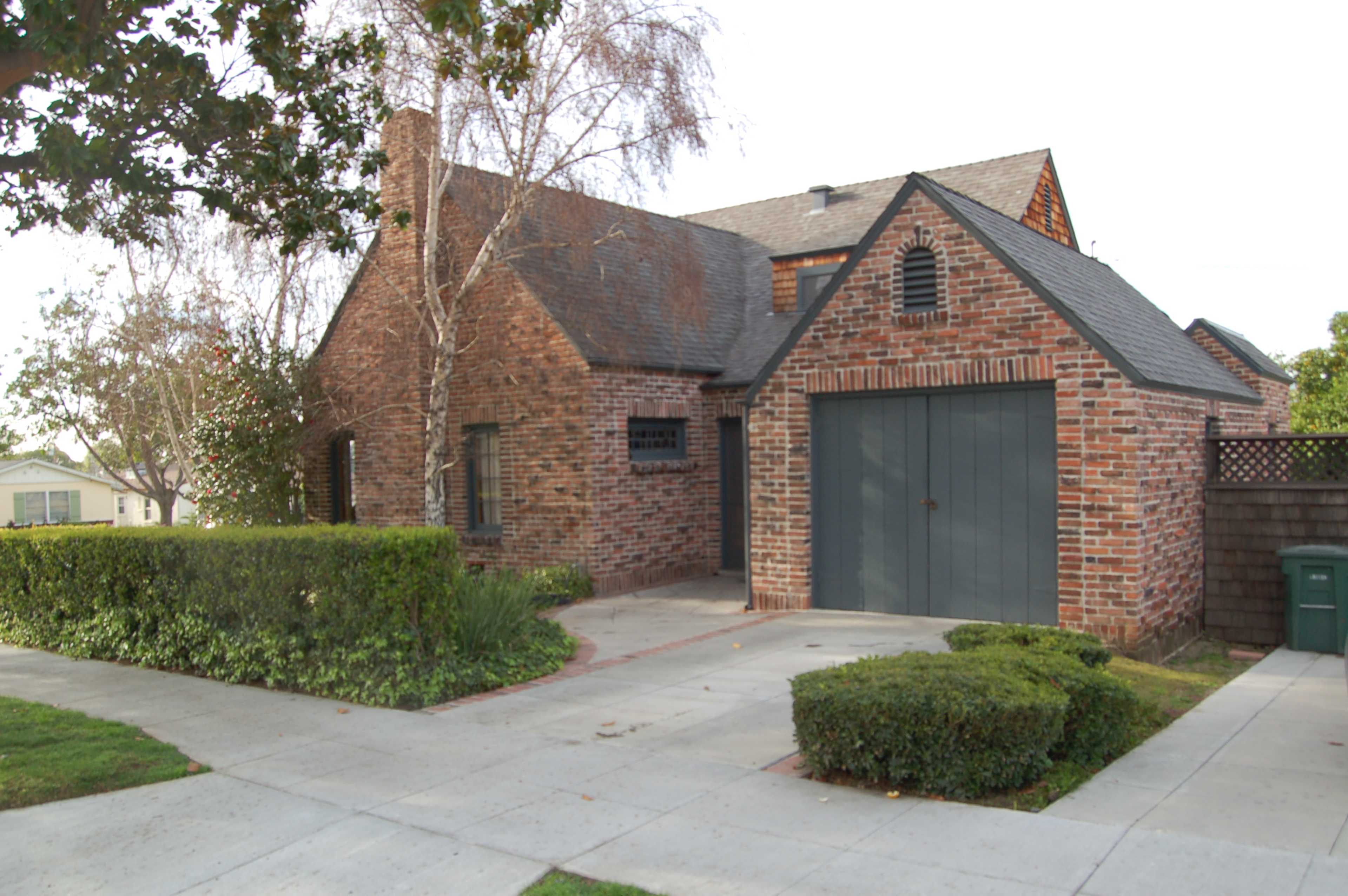 A brick house with a steep roof and a garage is situated on a residential street surrounded by neatly trimmed shrubs and a sidewalk.