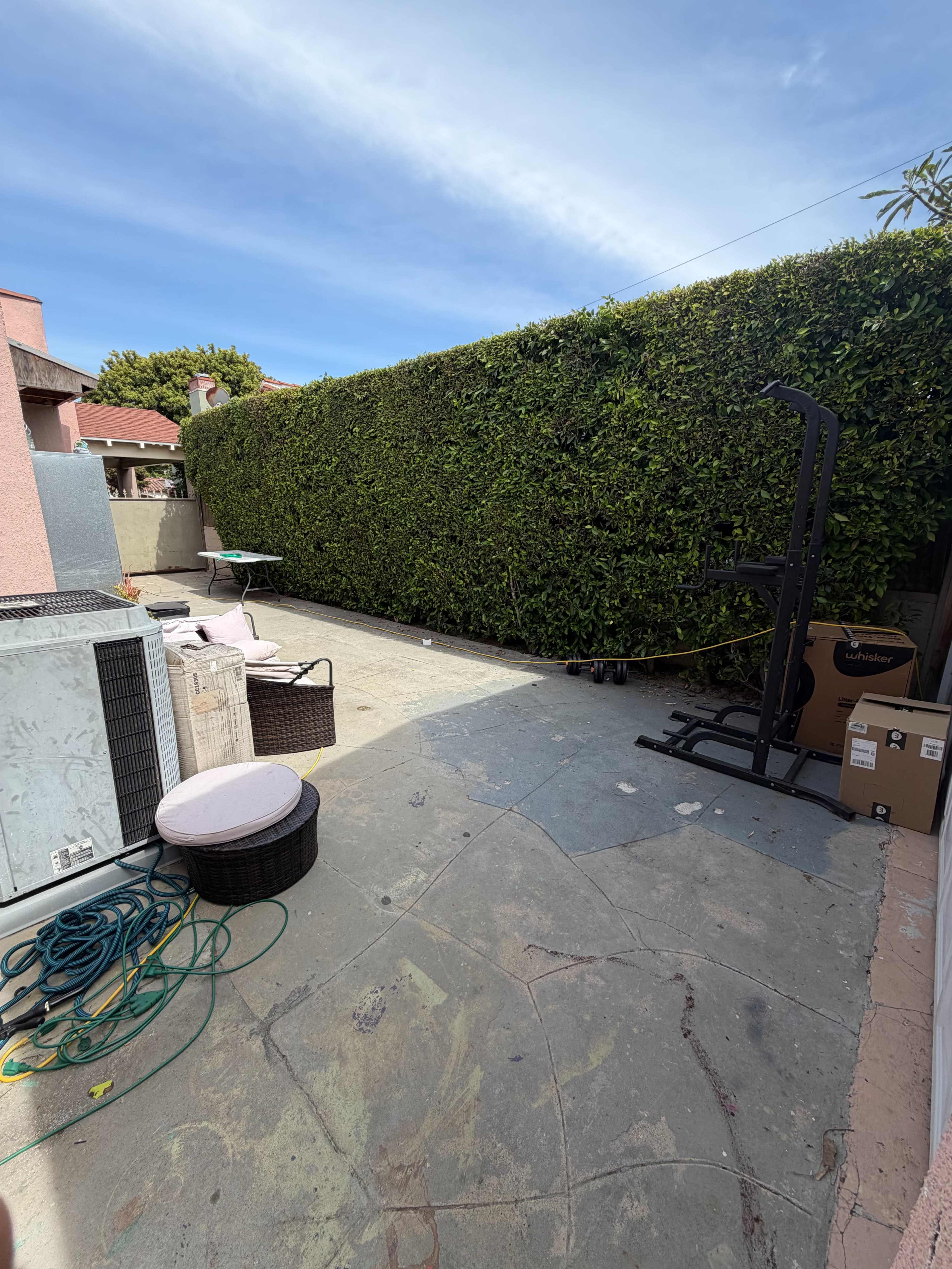 The image shows a fenced outdoor patio area with a concrete floor, some furniture items, and a backdrop of tall greenery.