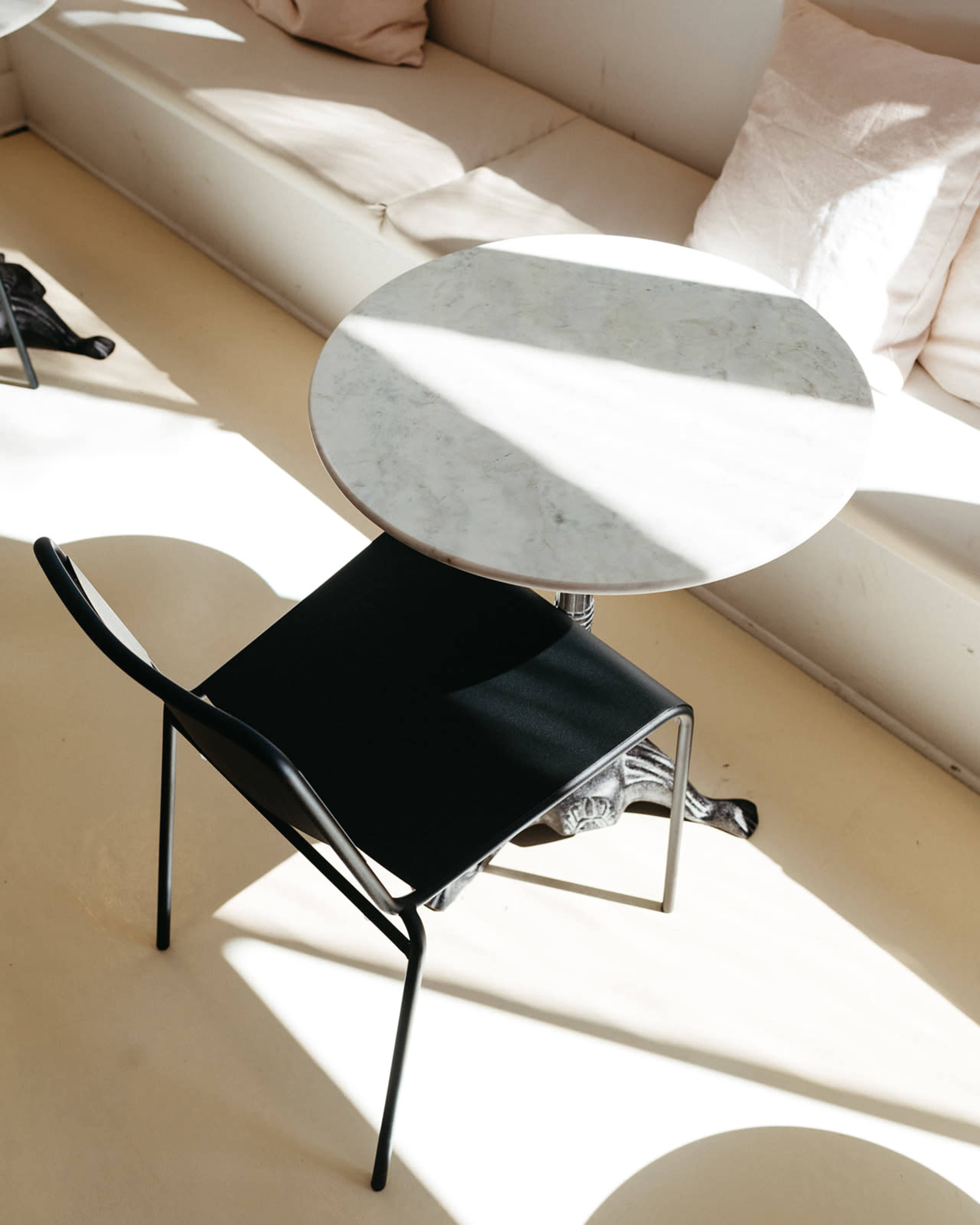 A round marble table sits next to a black metal chair on a light-colored floor, with sunlight casting shadows around them.