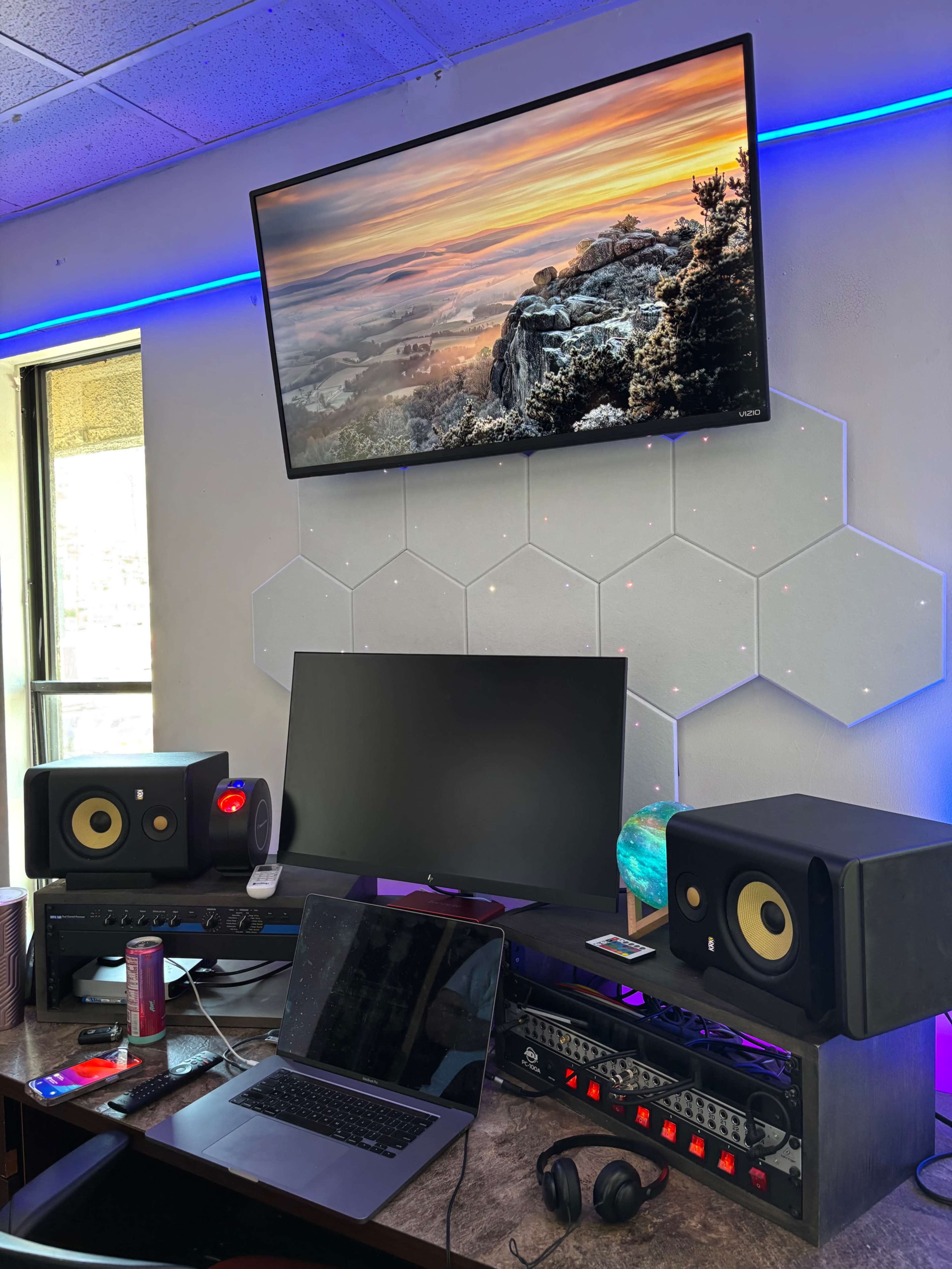 The image shows a modern workspace featuring a computer setup, studio monitors, a television displaying a landscape, and ambient lighting along the wall.