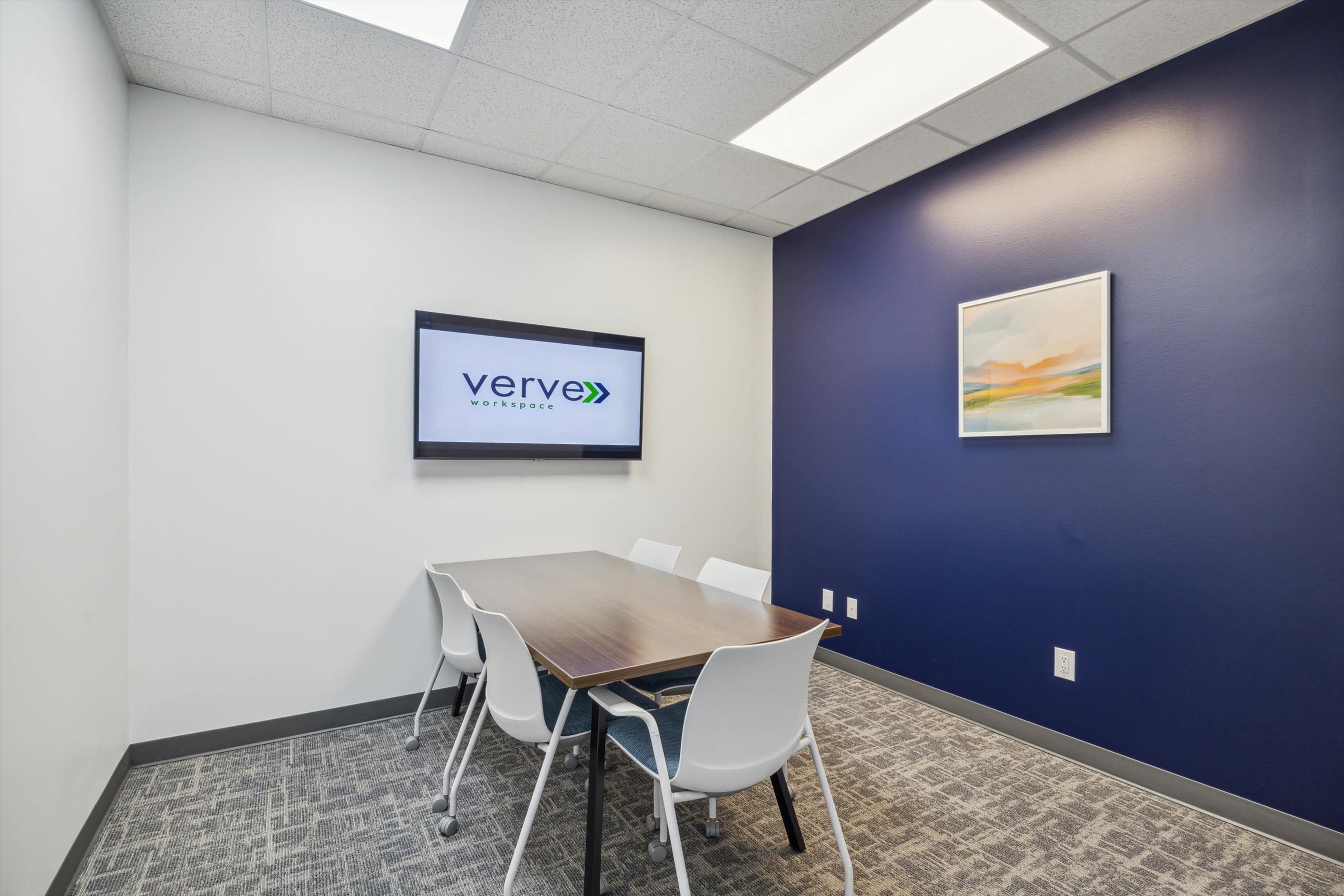 The image shows a small conference room with a brown table and four white chairs, featuring a wall-mounted screen displaying the "verve" logo.