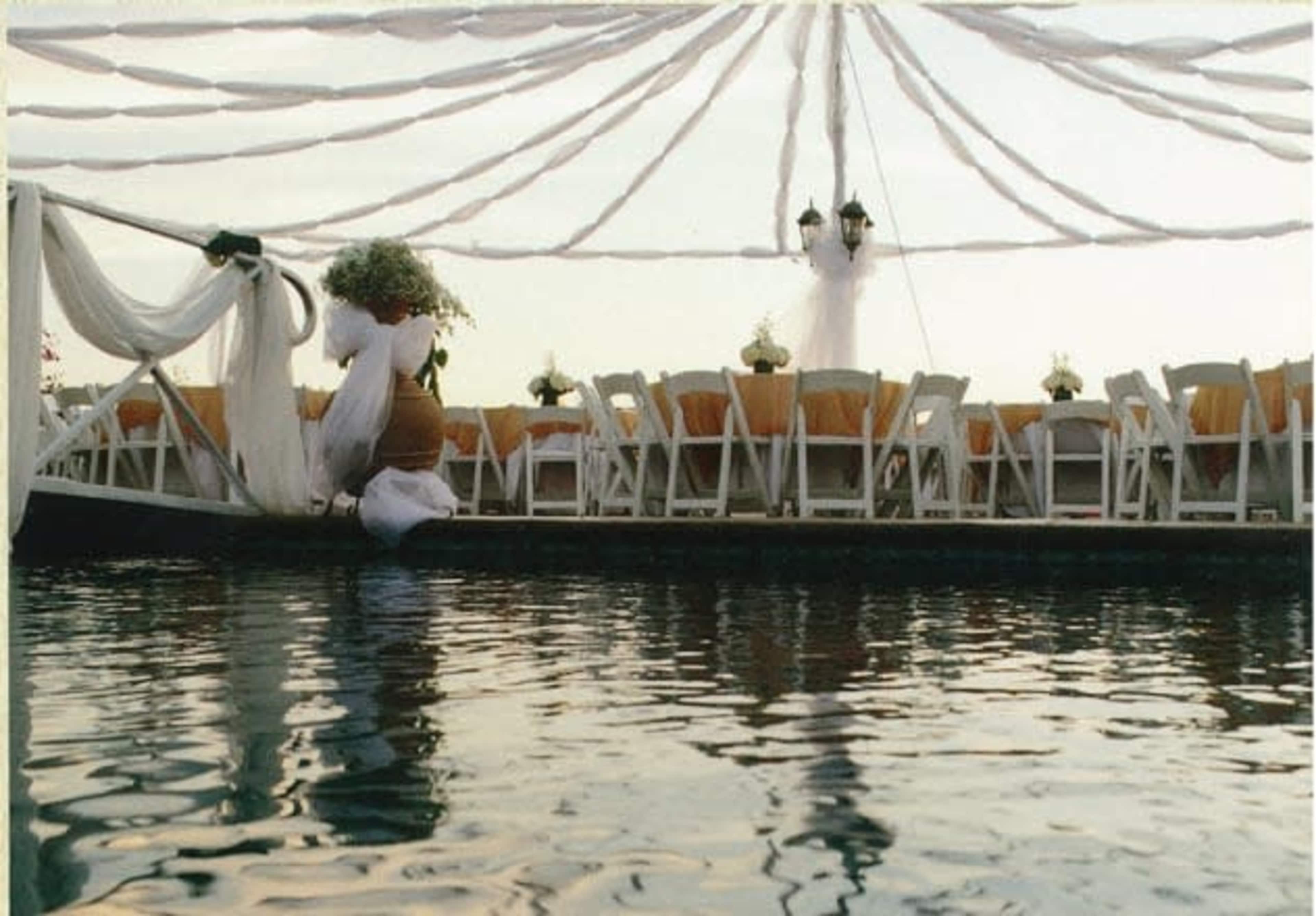 A decorated outdoor venue featuring a long table setting with chairs under a draped canopy, located over water.