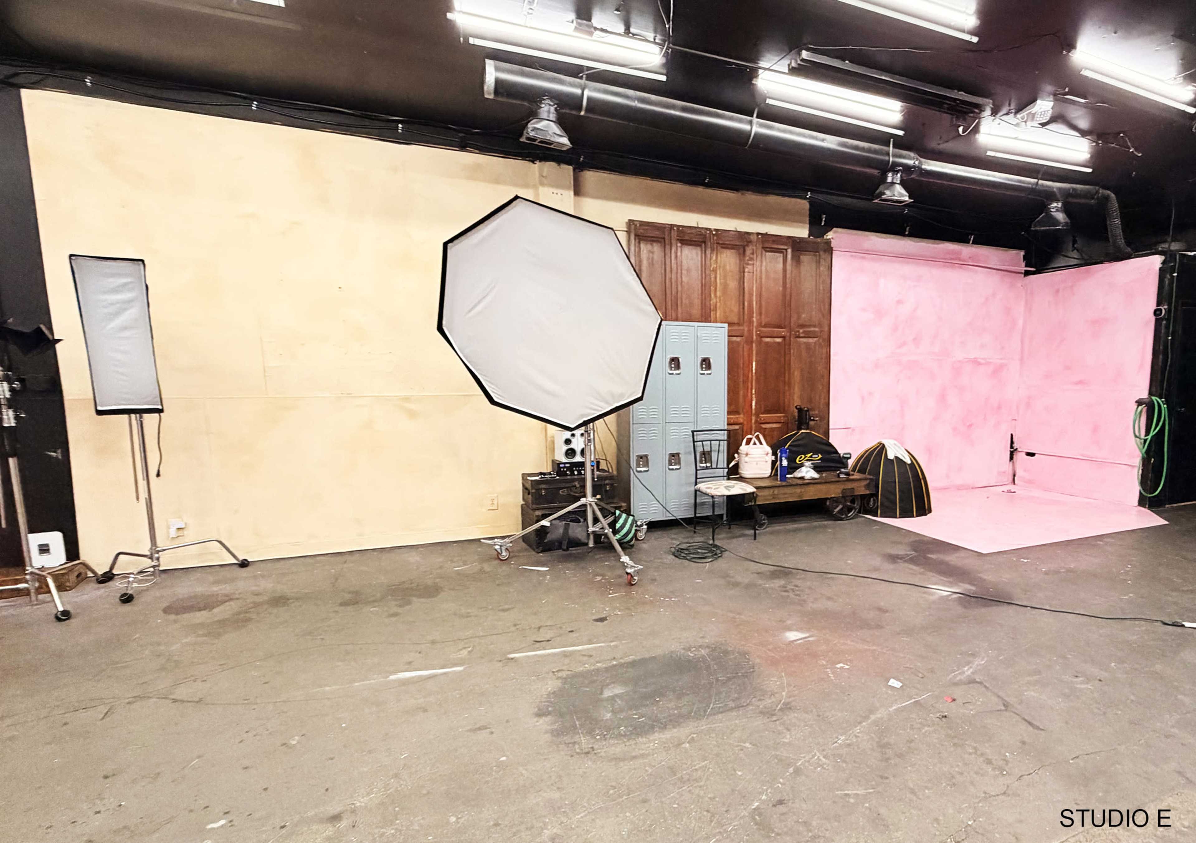 The image shows an industrial studio space featuring a pink backdrop, lighting equipment, and various props against a simple wall.