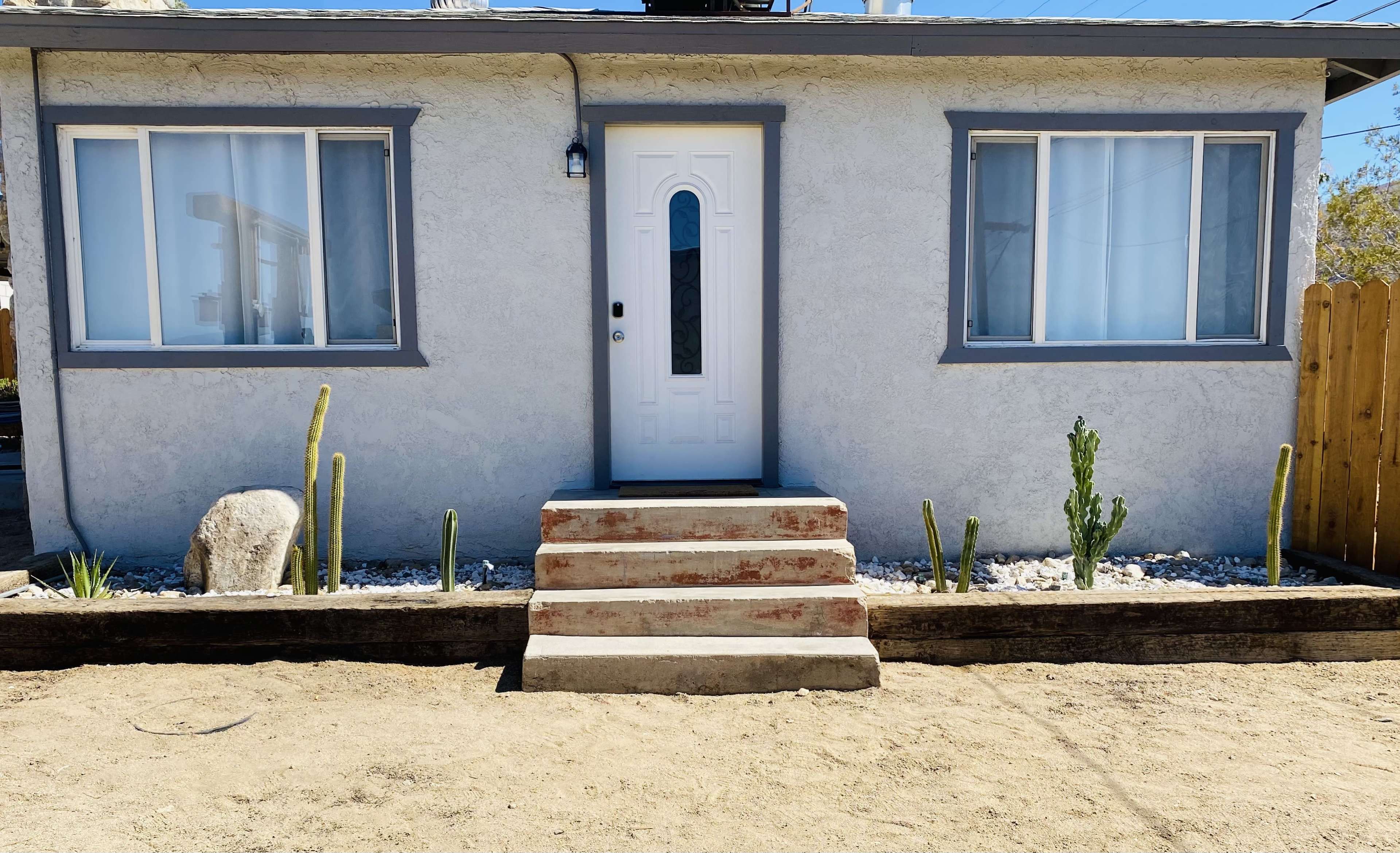 A small, light-colored house with a white door and two front windows is situated on a sandy area, featuring simple landscaping with cacti and gravel.