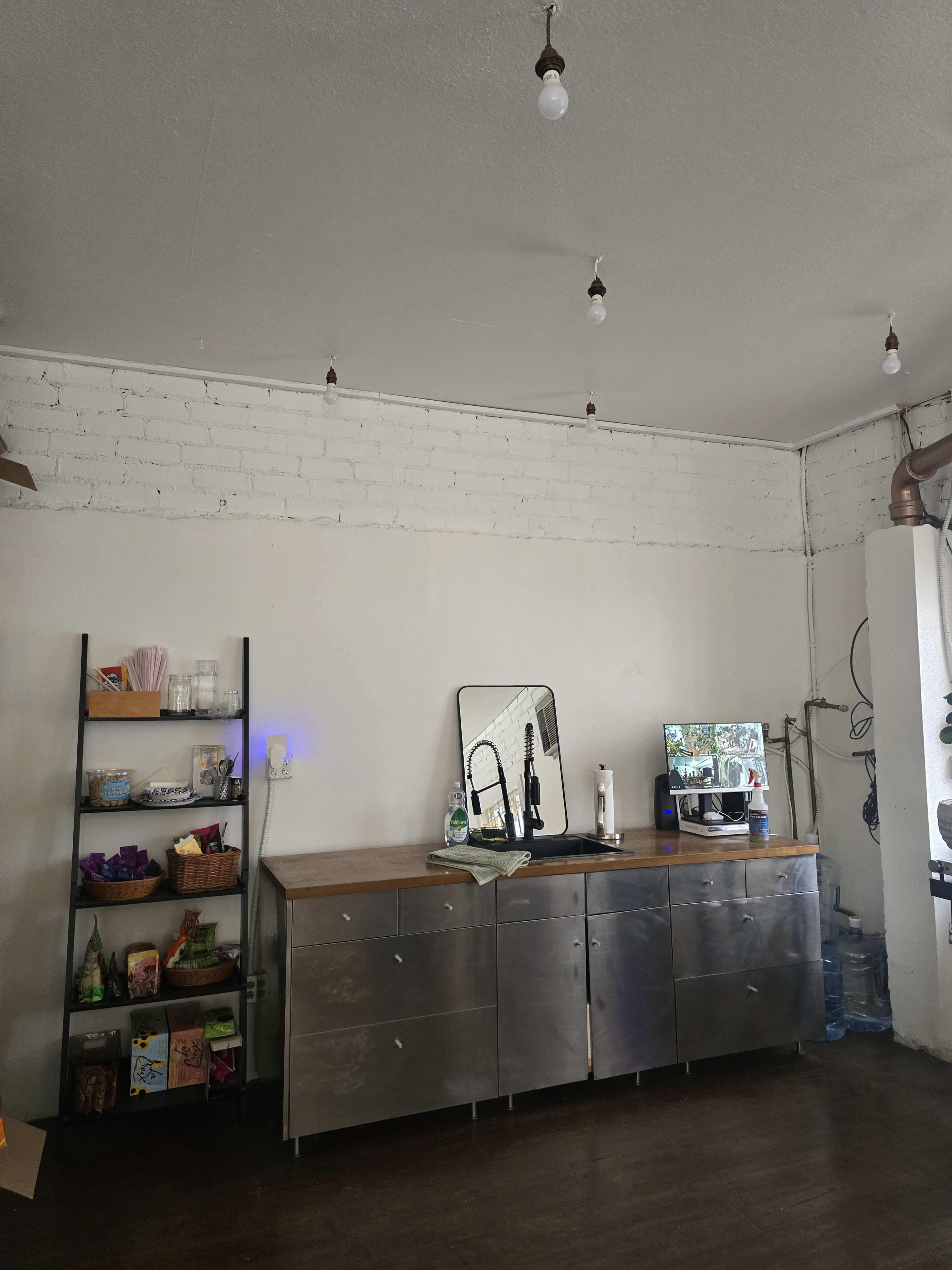 The image shows a simple indoor space featuring a metal cabinet below a mirror, a coffee machine, and shelves filled with various items.