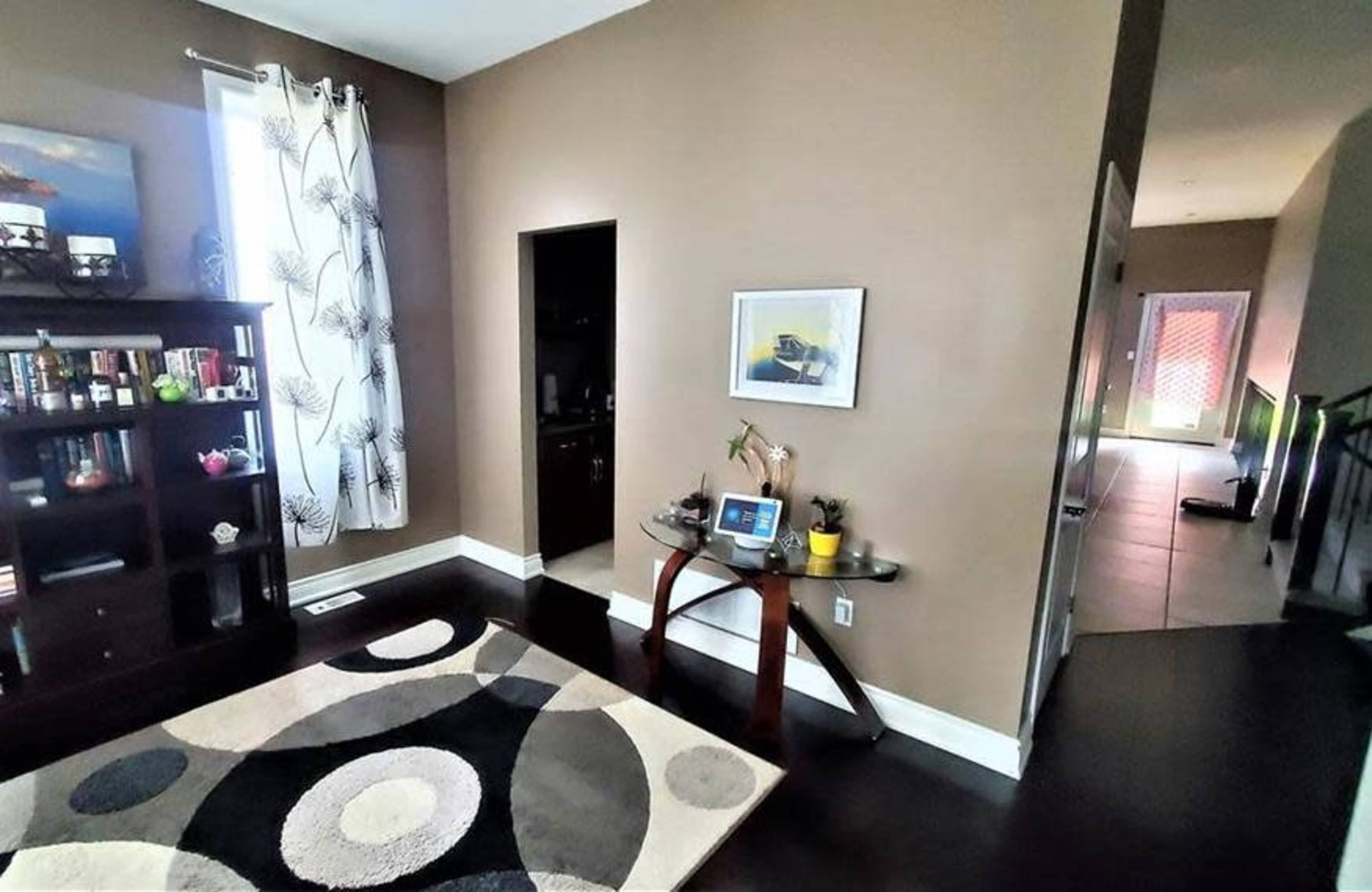 A living room with dark hardwood flooring, a decorative area rug, a bookshelf, and a small table with electronic devices, leading to a hallway and another room visible in the background.