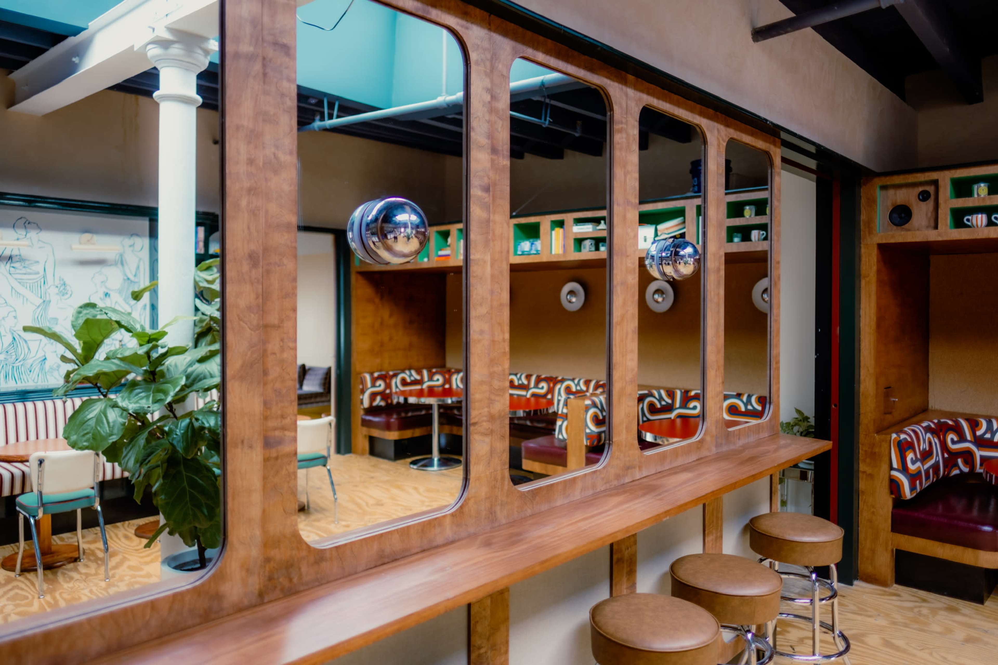 The image shows a stylish, modern interior featuring a wooden bar counter, three stools, and a series of tall mirrors reflecting the space.