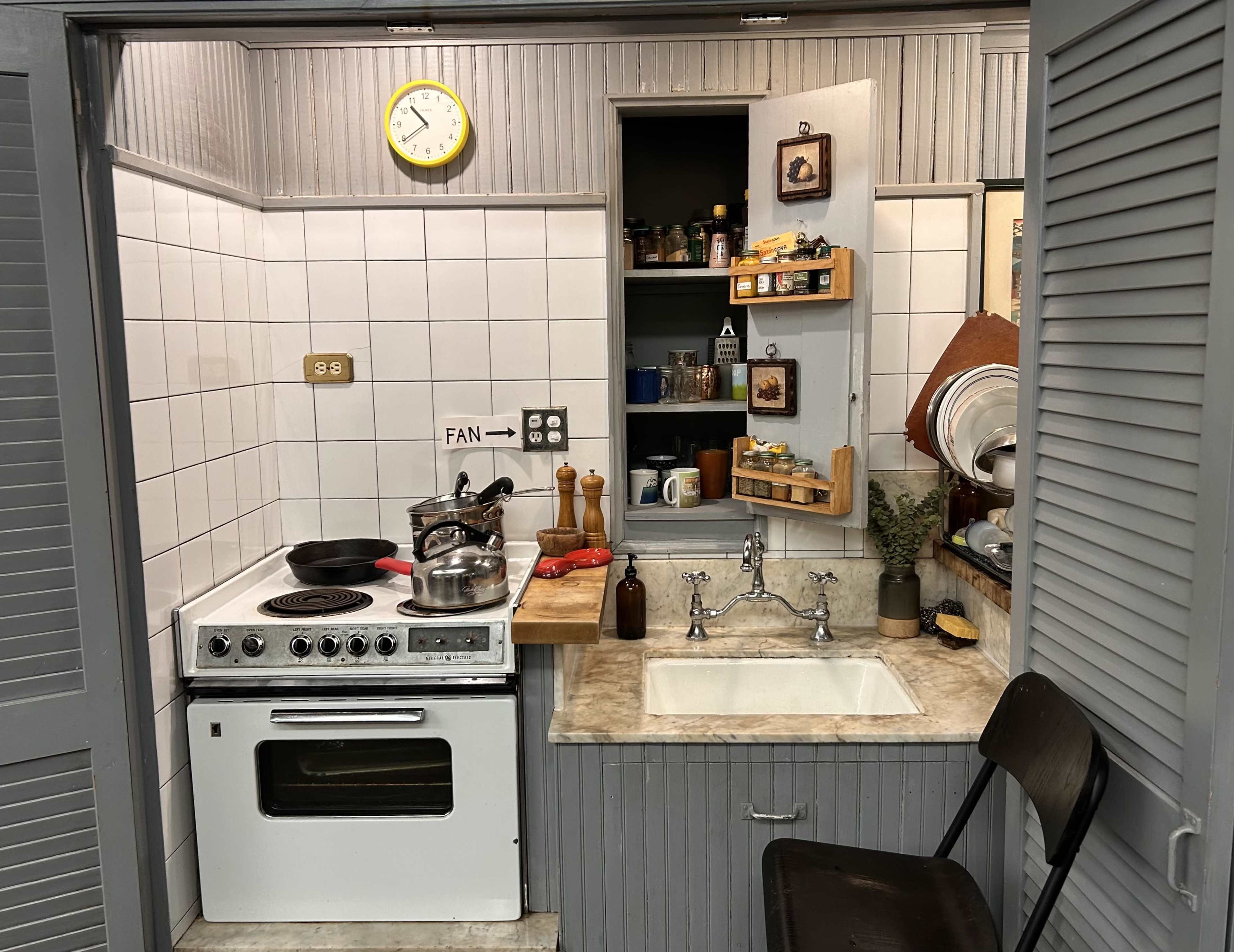 The image shows a compact kitchen with a stove, sink, wooden countertop, and shelves filled with various containers and utensils.