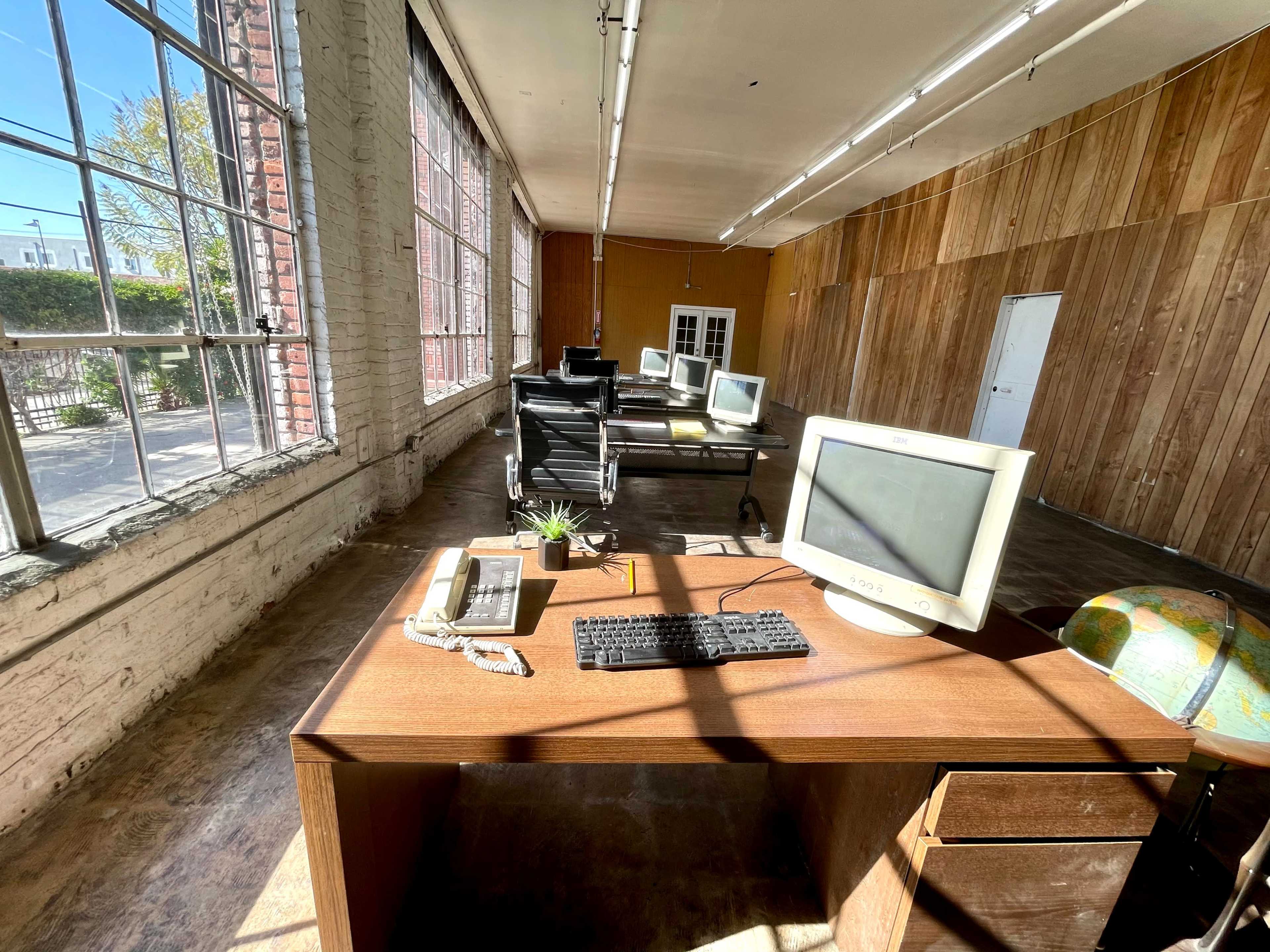 An empty office space features a wooden desk with a computer, telephone, and globe, while several unused computer monitors are lined up in the background near large windows.