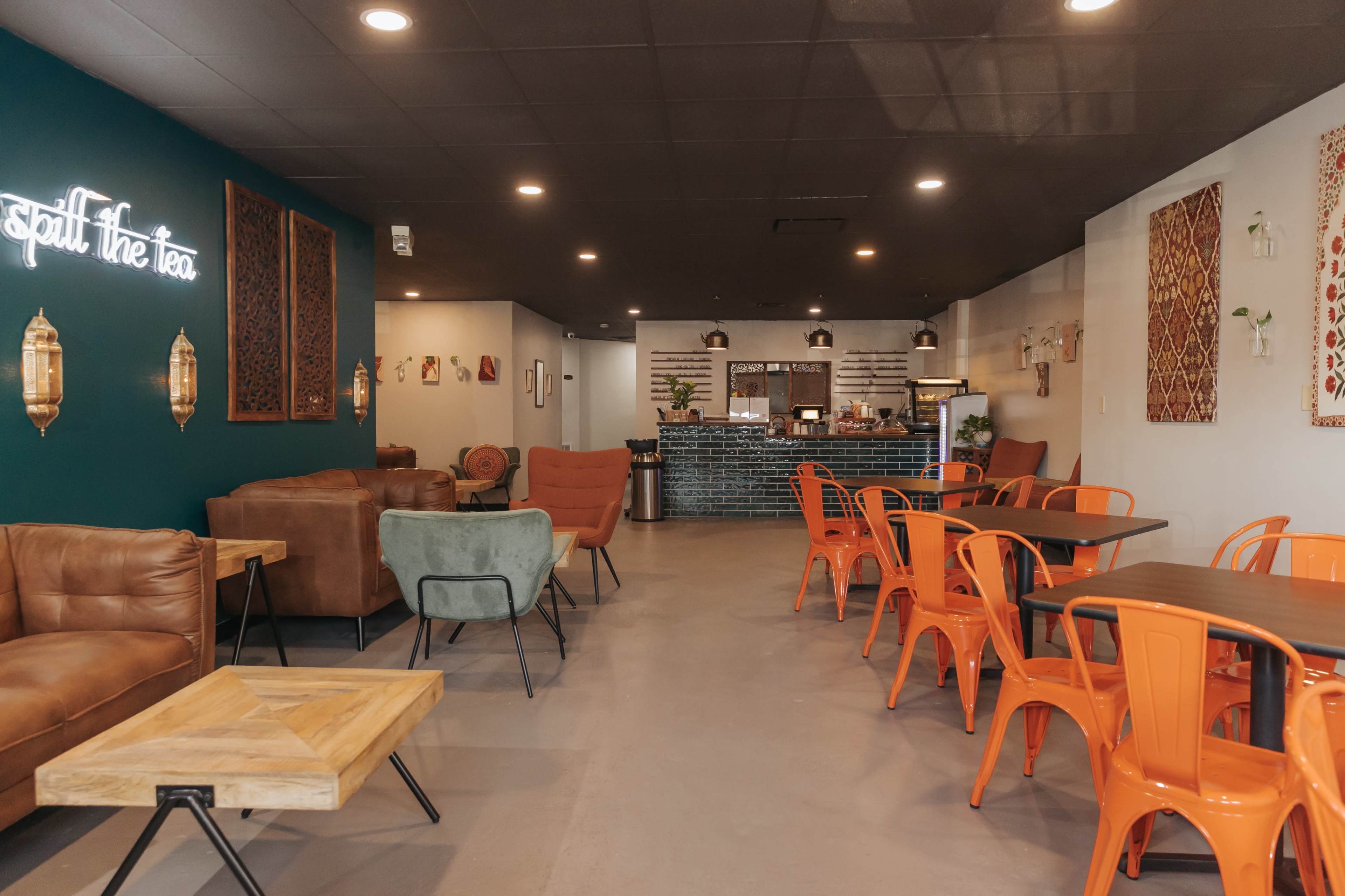 A café interior featuring orange chairs, a wooden table, and a green wall with neon signage, along with a service counter in the background.